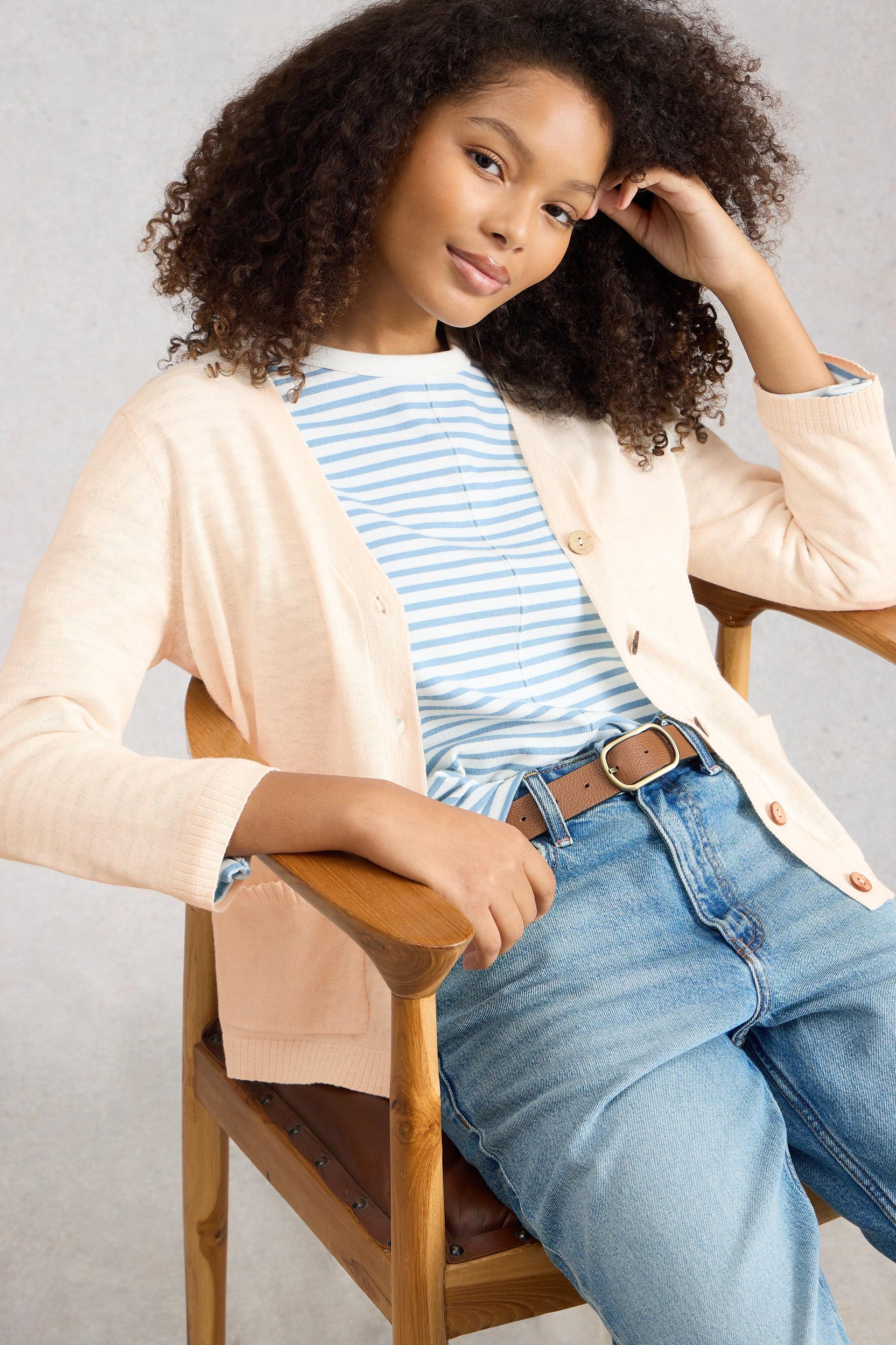 Model sitting on a chair wearing a striped shirt, beige organic cotton linen cardigan, and blue jeans.