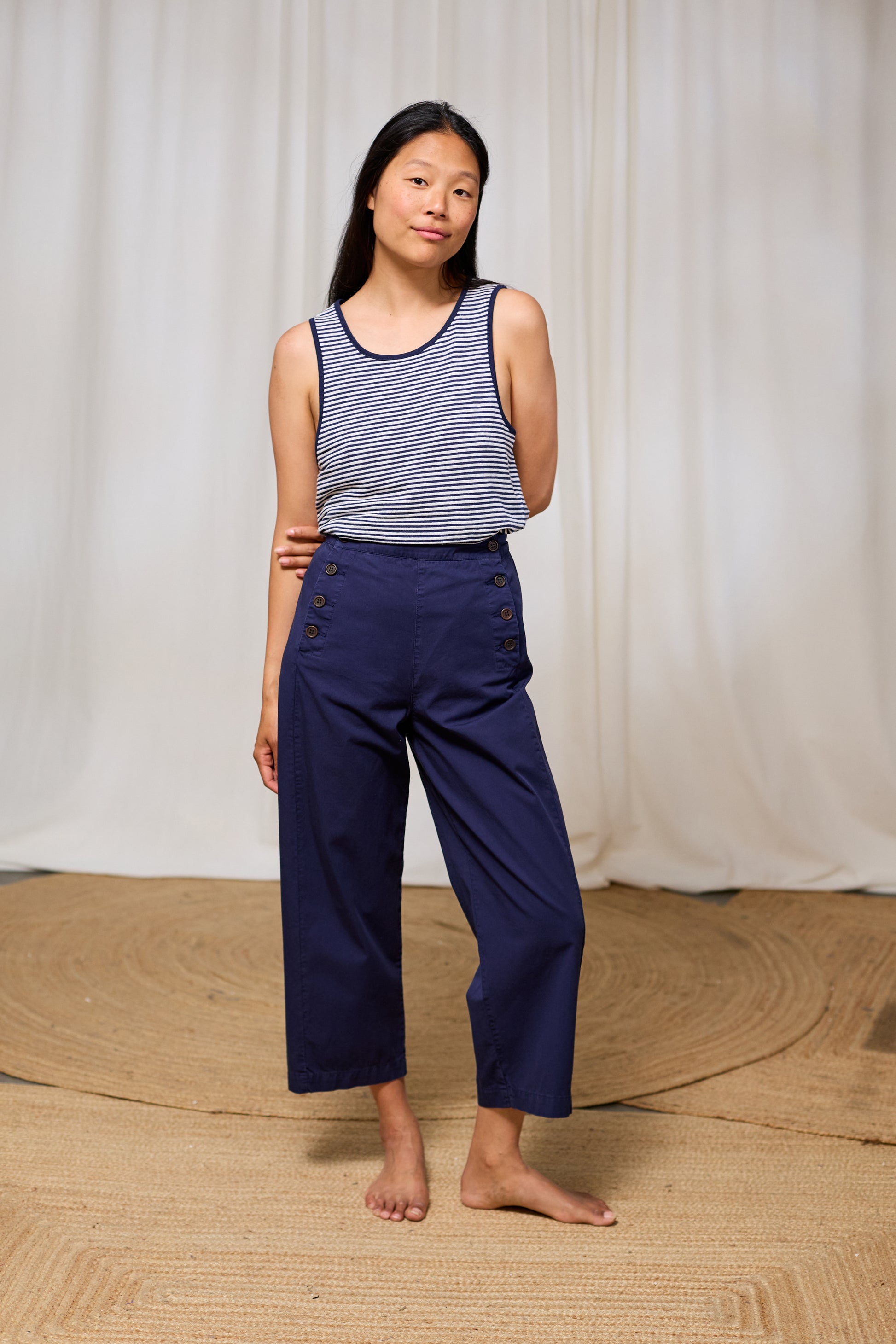 Woman wearing a striped tank top and navy pants standing against a white curtain.