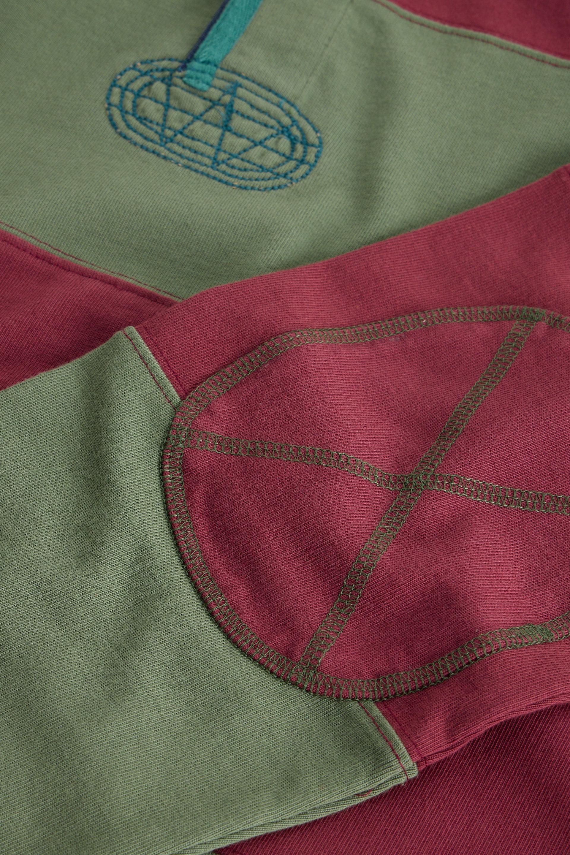 Close-up of a patchwork fabric garment with sage green and dusty rose sections, featuring contrasting brown stitching and a blue embroidered logo.
