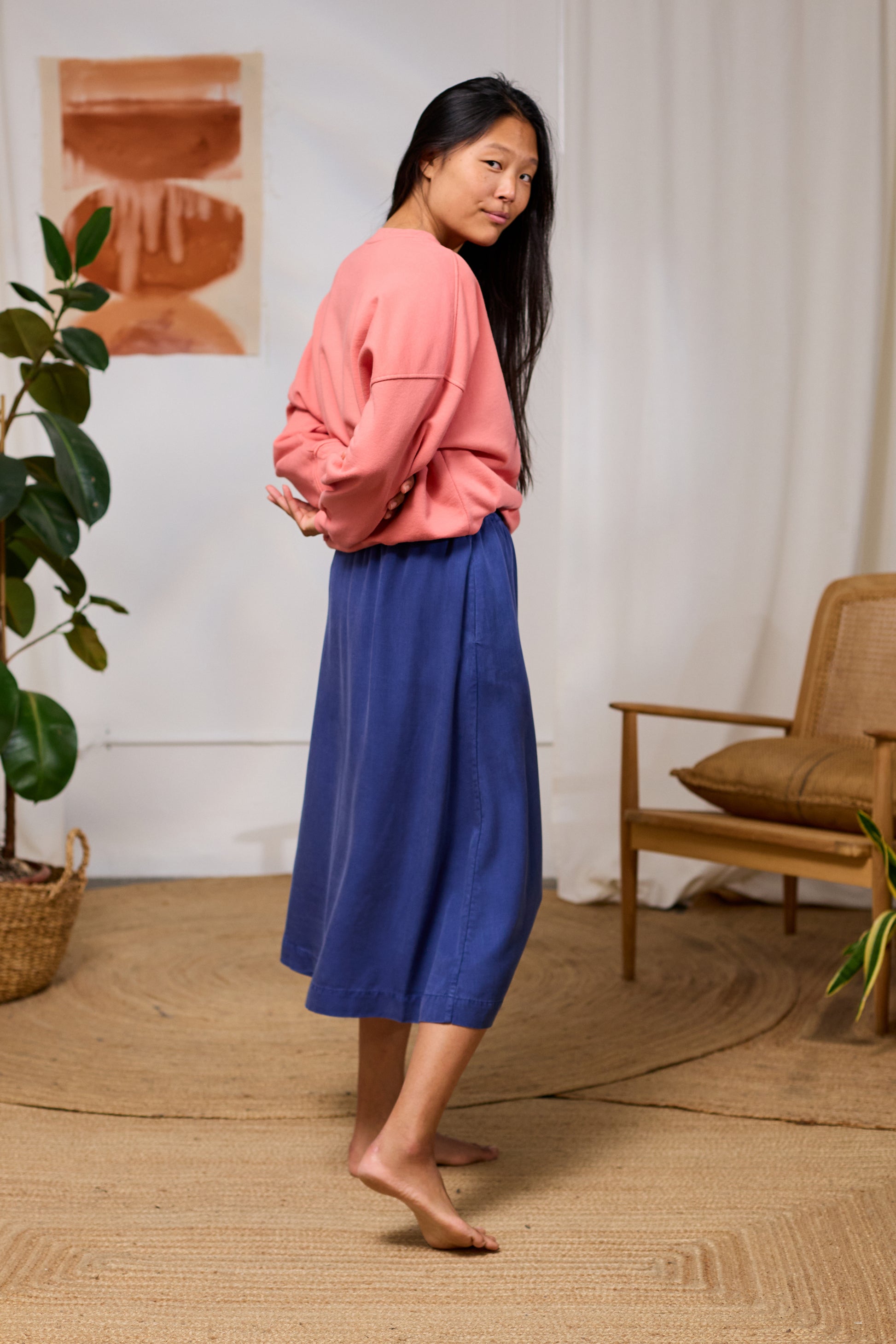 Woman in pink TWOTHIRDS sweater and blue skirt standing in a room with a plant and chair.