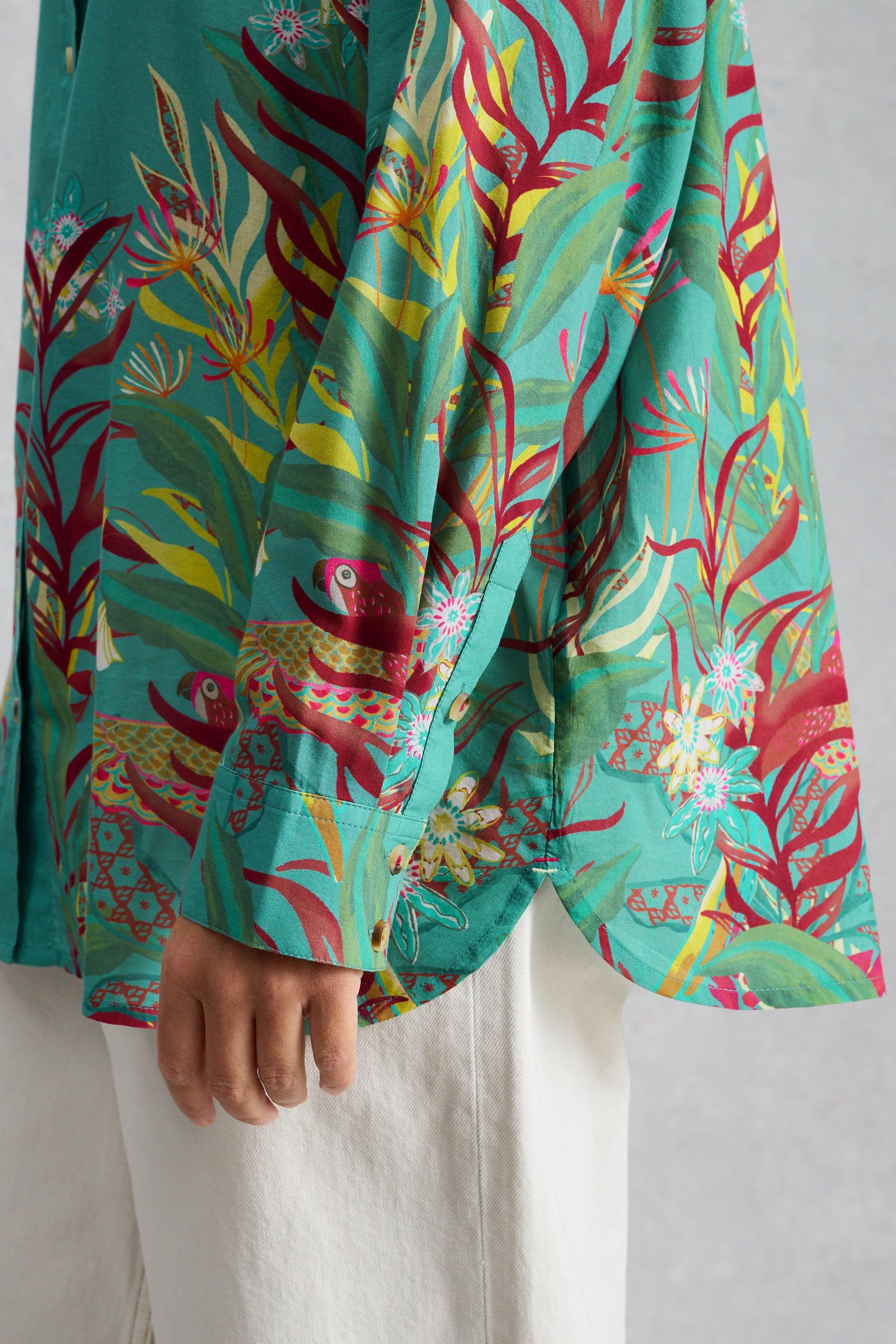 Close-up of a person wearing a vibrant teal organic cotton printed shirt with parrot and floral patterns.