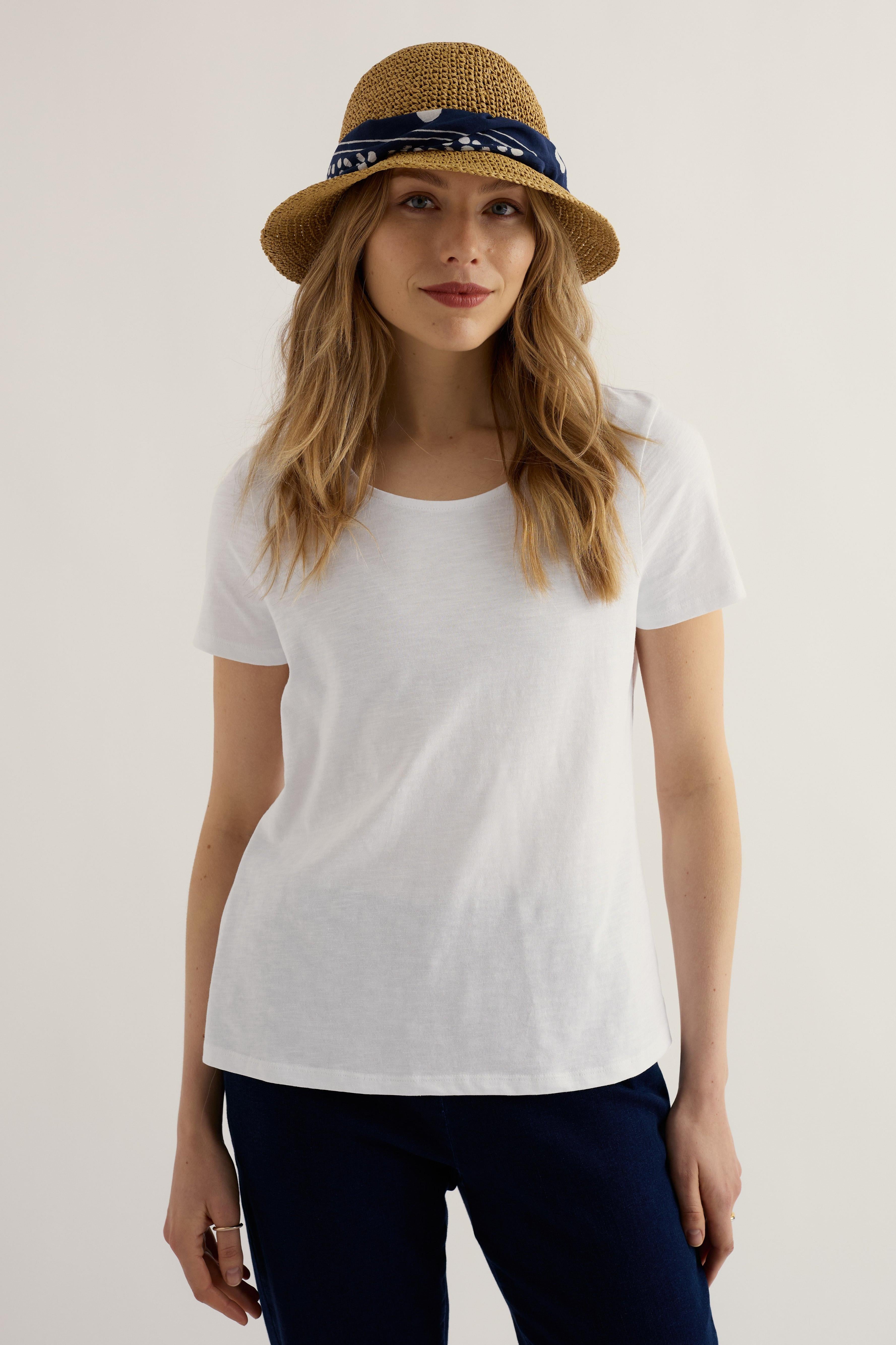Woman wearing a white t-shirt, navy pants, and a straw hat with a bandana on a plain background