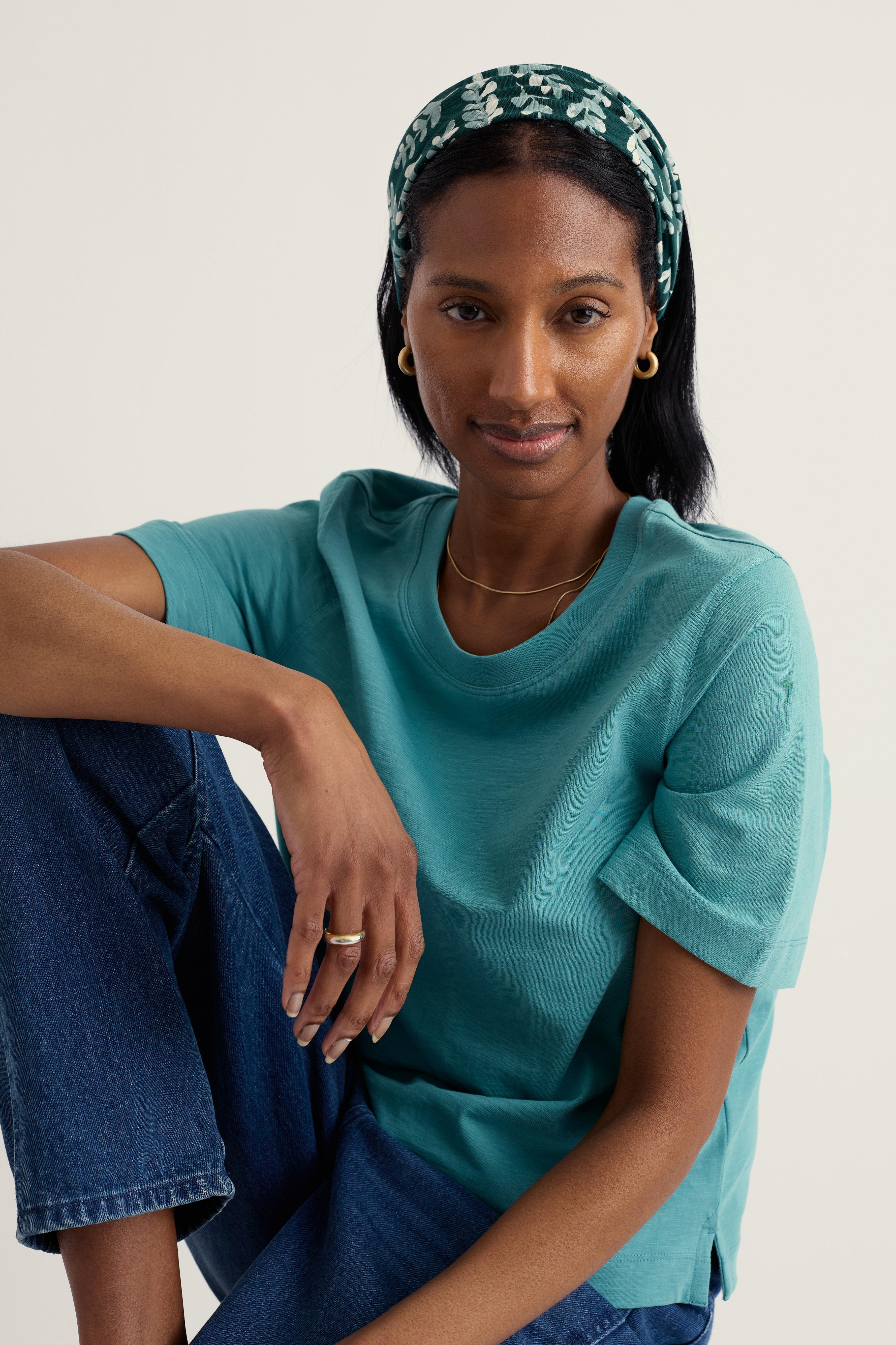 Woman wearing a teal shirt and blue jeans with a patterned Seasalt handyband on a plain background