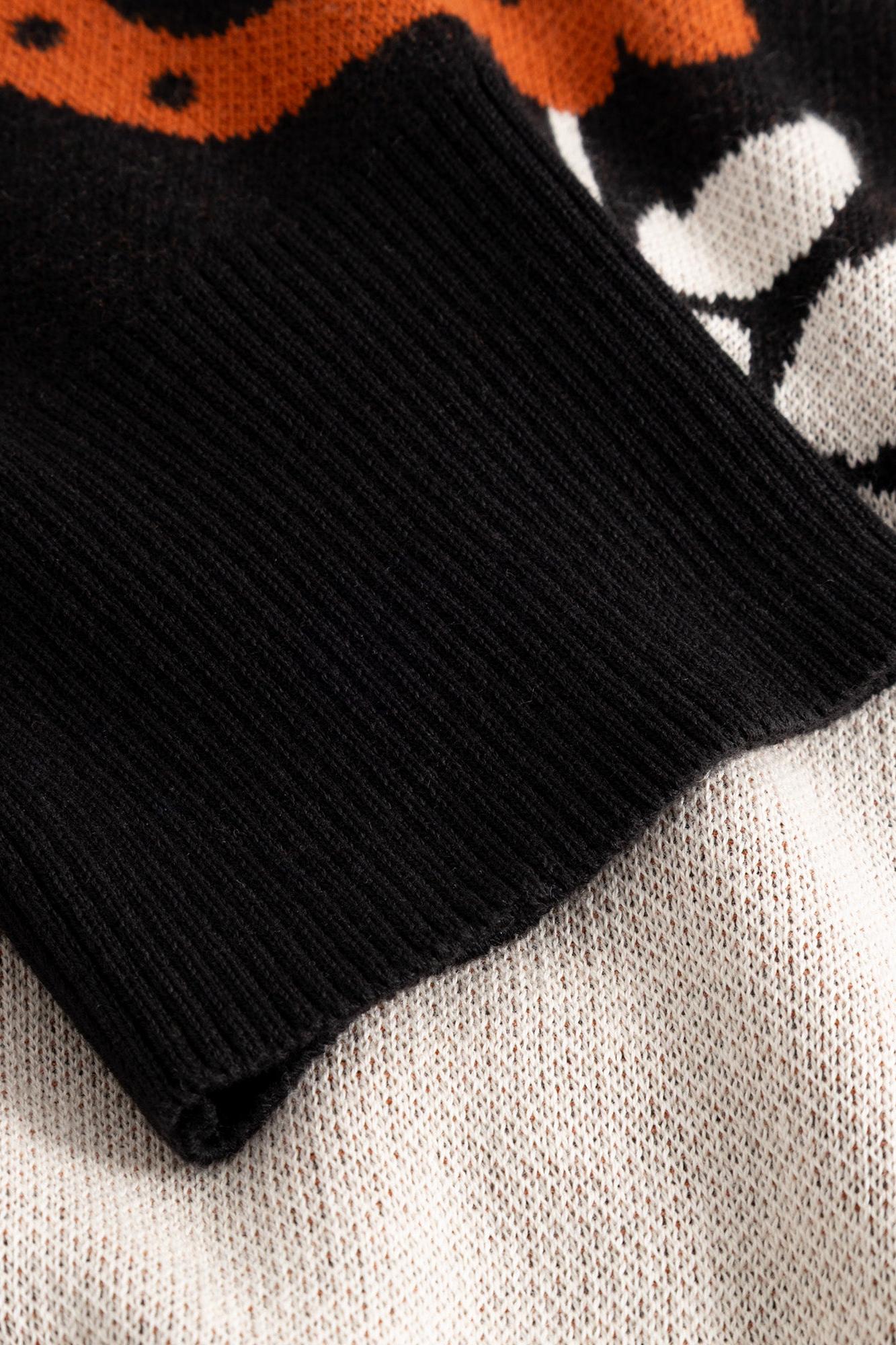 Close-up of a knit sweater cuff in solid black ribbed knit, layered over a cream, orange, and black patterned knit body.