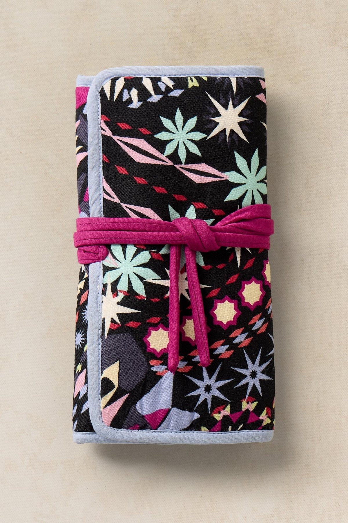 A folded, patterned fabric jewellery organiser with a magenta ribbon tie closure on a neutral background.