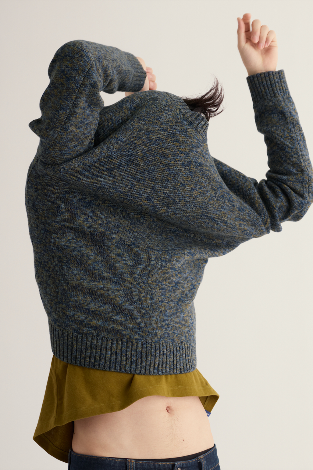 A person wearing a loose, oversized knit sweater in a heathered blue-gray and olive tone, shown from the back against a plain light beige studio background.