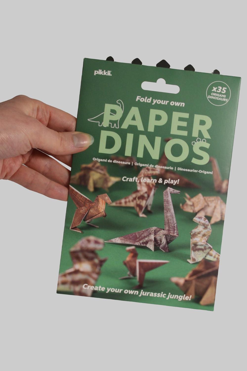 Hand holding a green package of 'Fold your own Paper Dinos' craft kit on a gray background