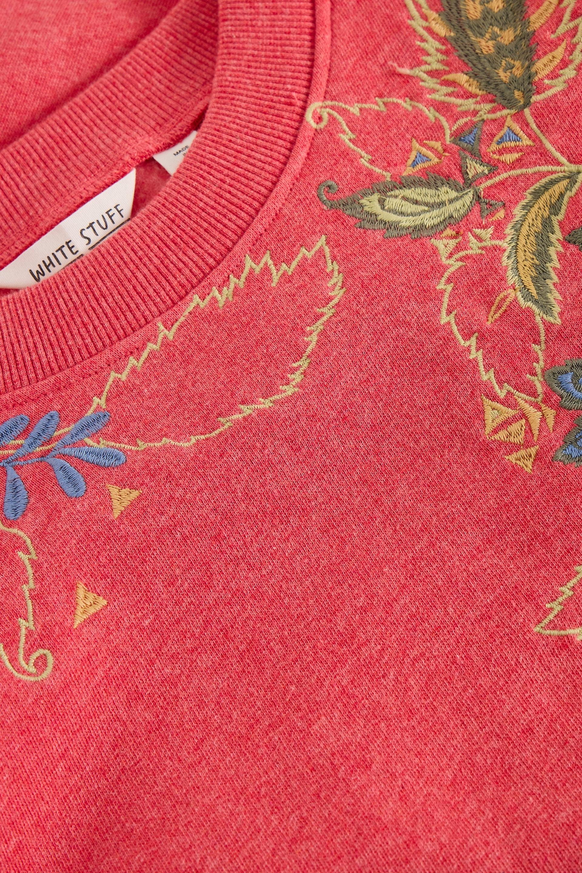 Close-up of a coral pink crewneck sweatshirt with a colorful embroidered floral pattern in yellow, green, and blue. A white neck label reads 'WHITE STUFF'.