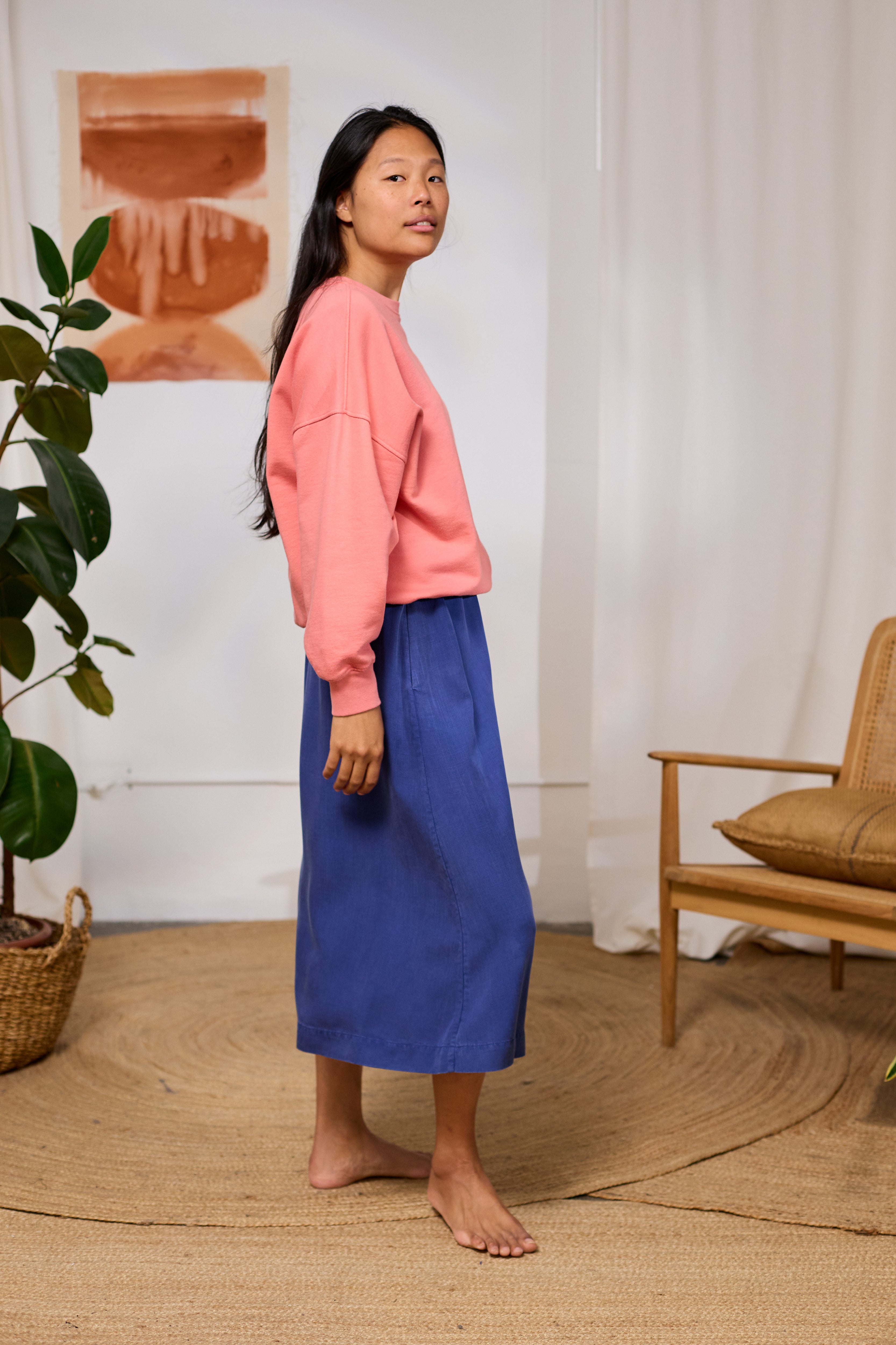 Woman in pink TWOTHIRDS sweater and blue skirt standing in a room with a plant and chair.