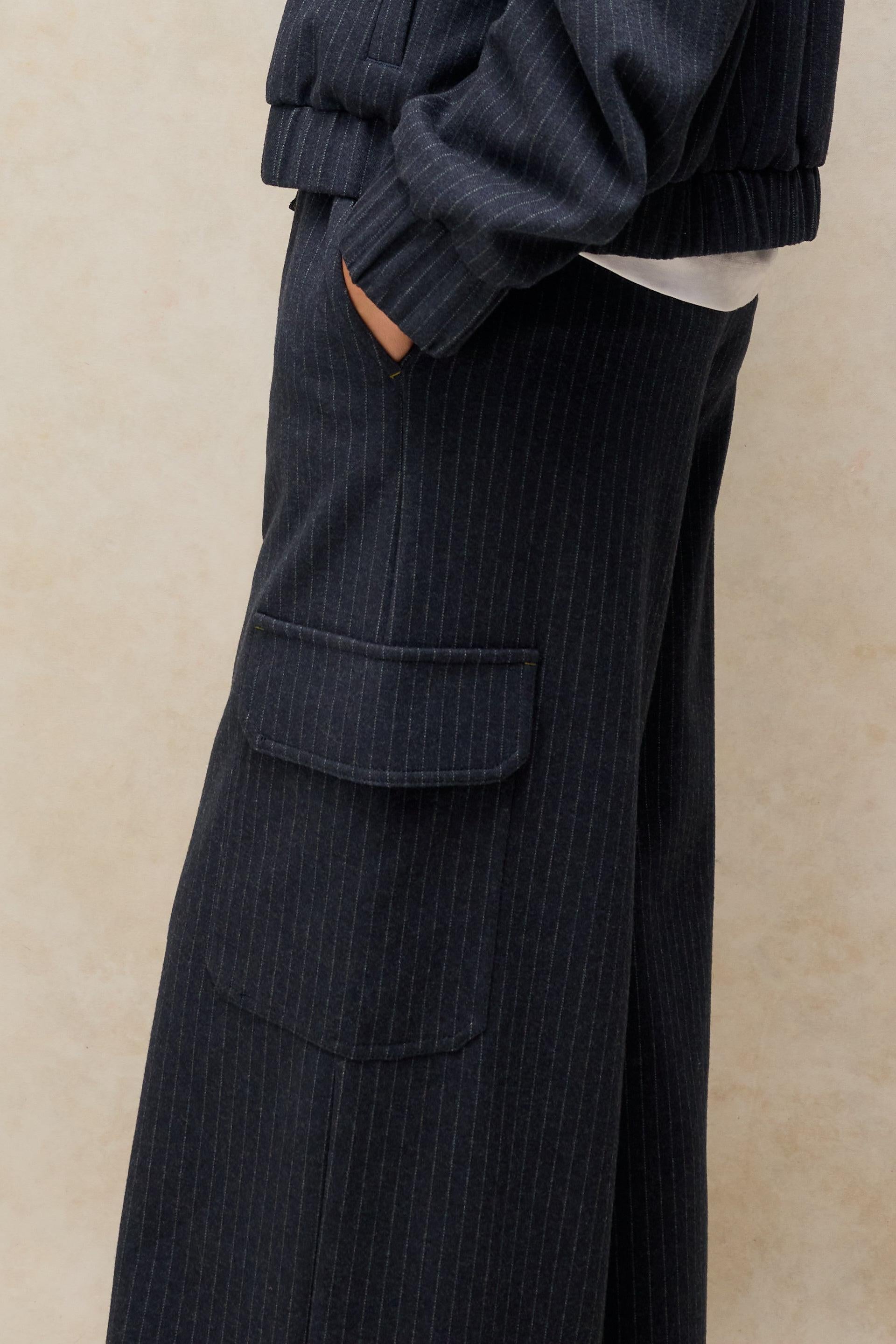 Close-up of a person wearing dark navy pinstriped wide-leg trousers with a large front flap pocket, paired with a matching dark ribbed top against a warm beige textured background.