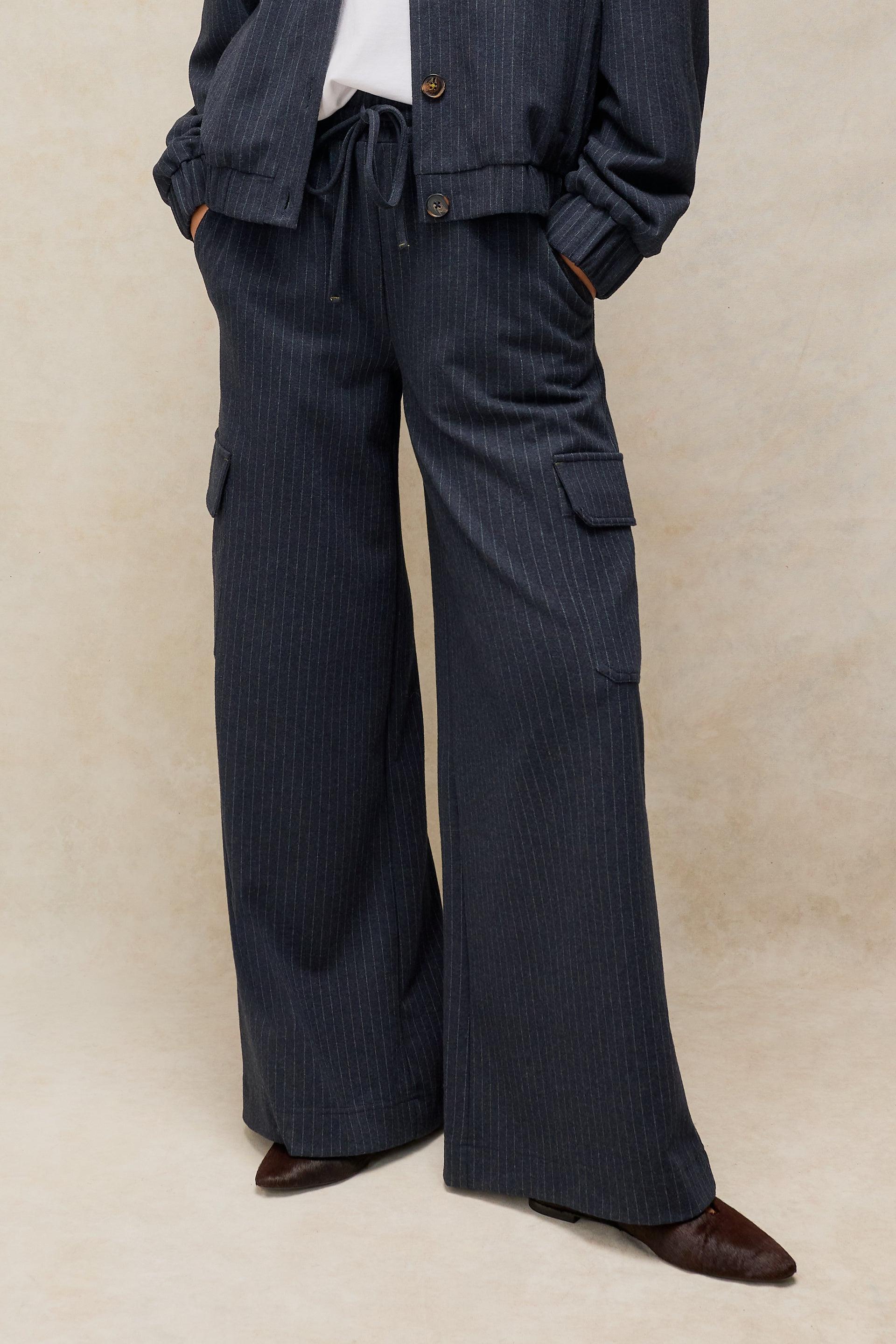 A person wearing dark navy pinstriped wide-leg trousers with cargo pockets, paired with a matching jacket and brown pointed-toe shoes, against a neutral beige studio backdrop.