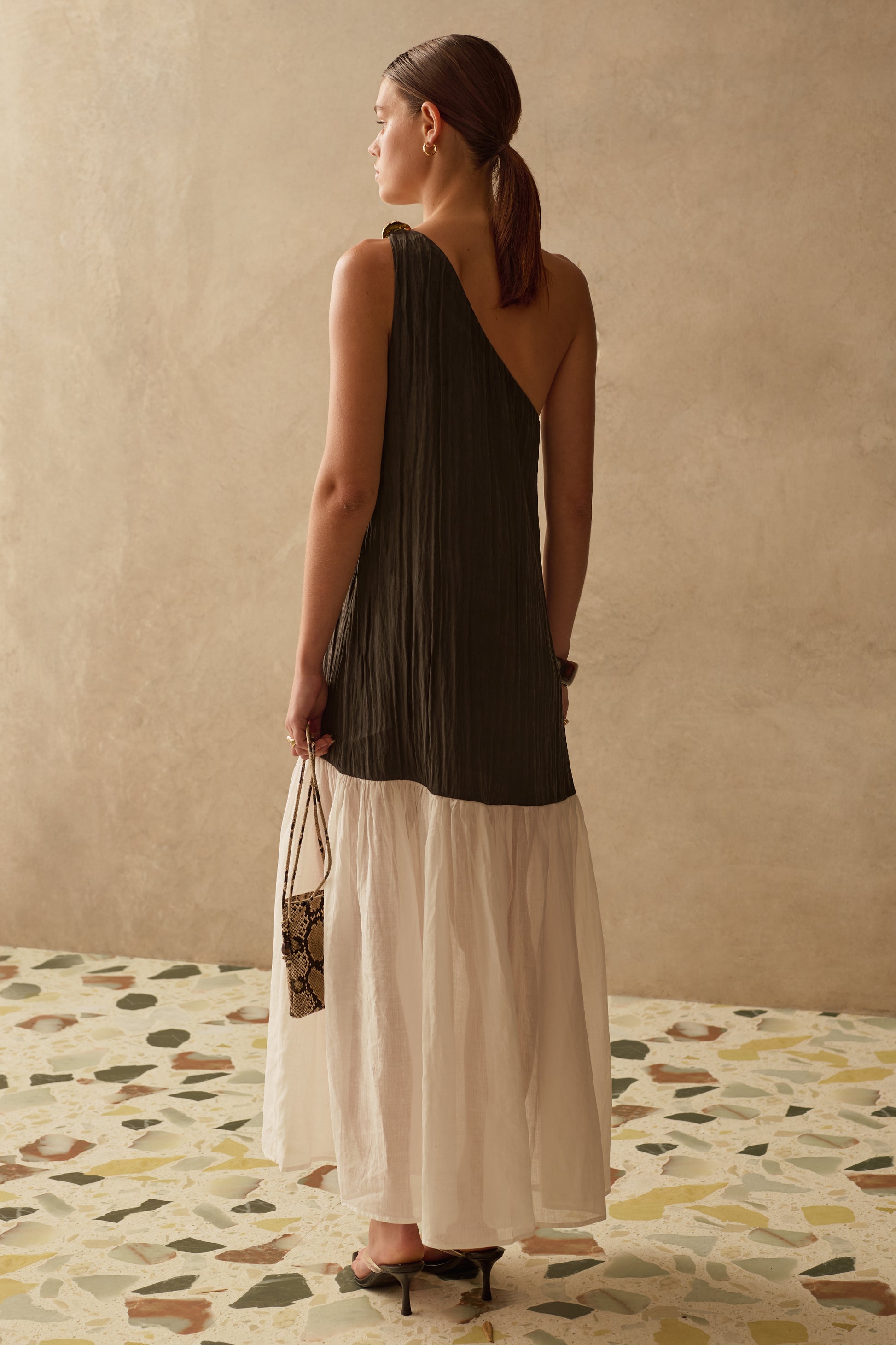 Woman wearing a black asymmetric maxi dress standing against a beige wall with a patterned floor.