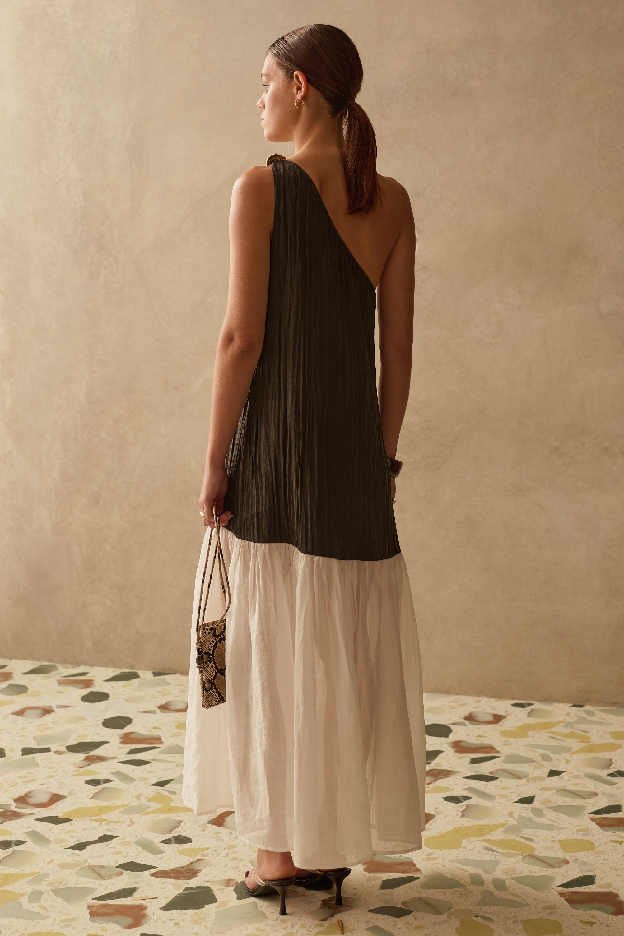 Woman wearing a black asymmetric maxi dress standing against a beige wall with a patterned floor.
