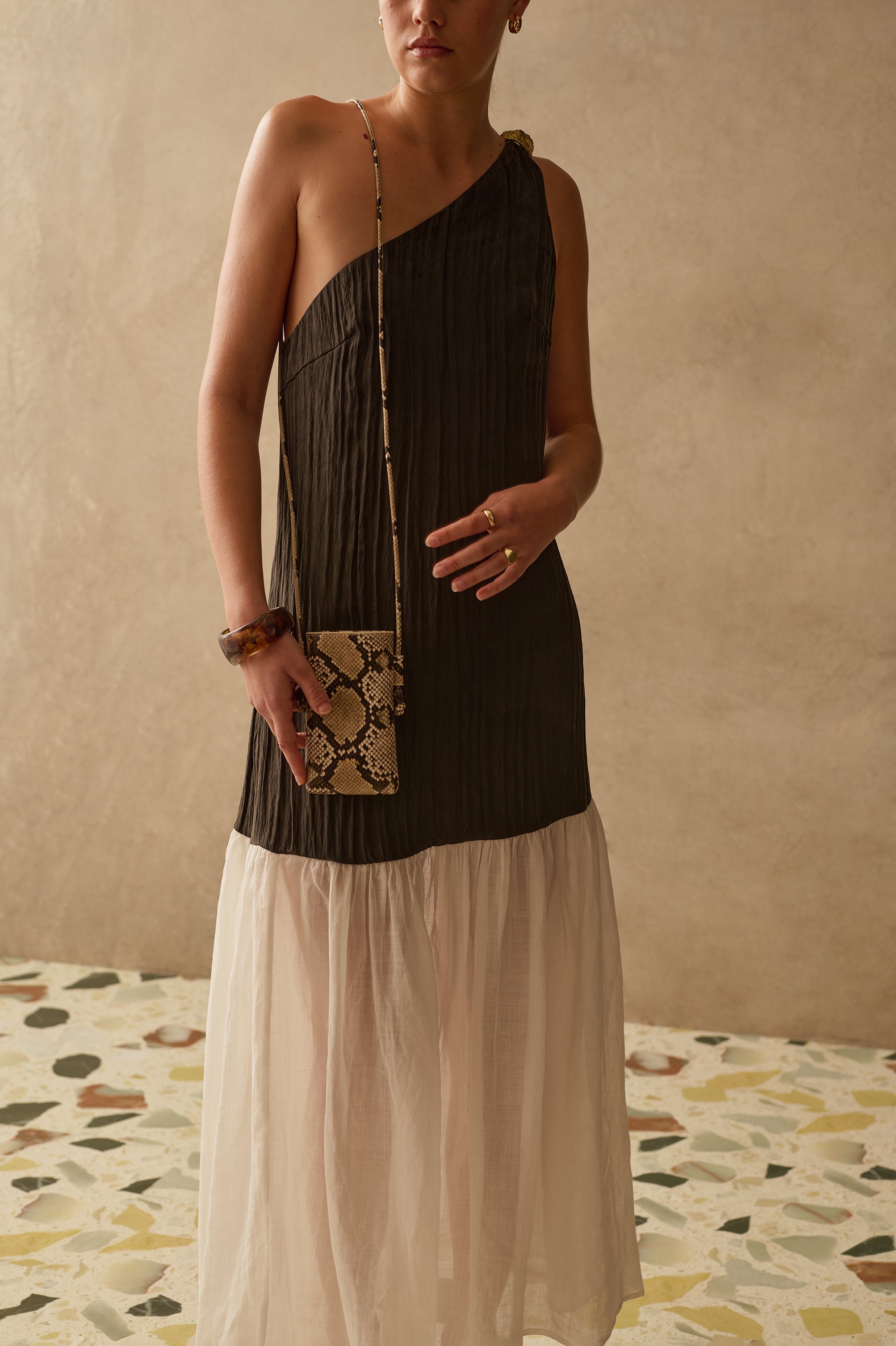 Woman wearing a black one-shoulder wedding guest dress against a beige wall.