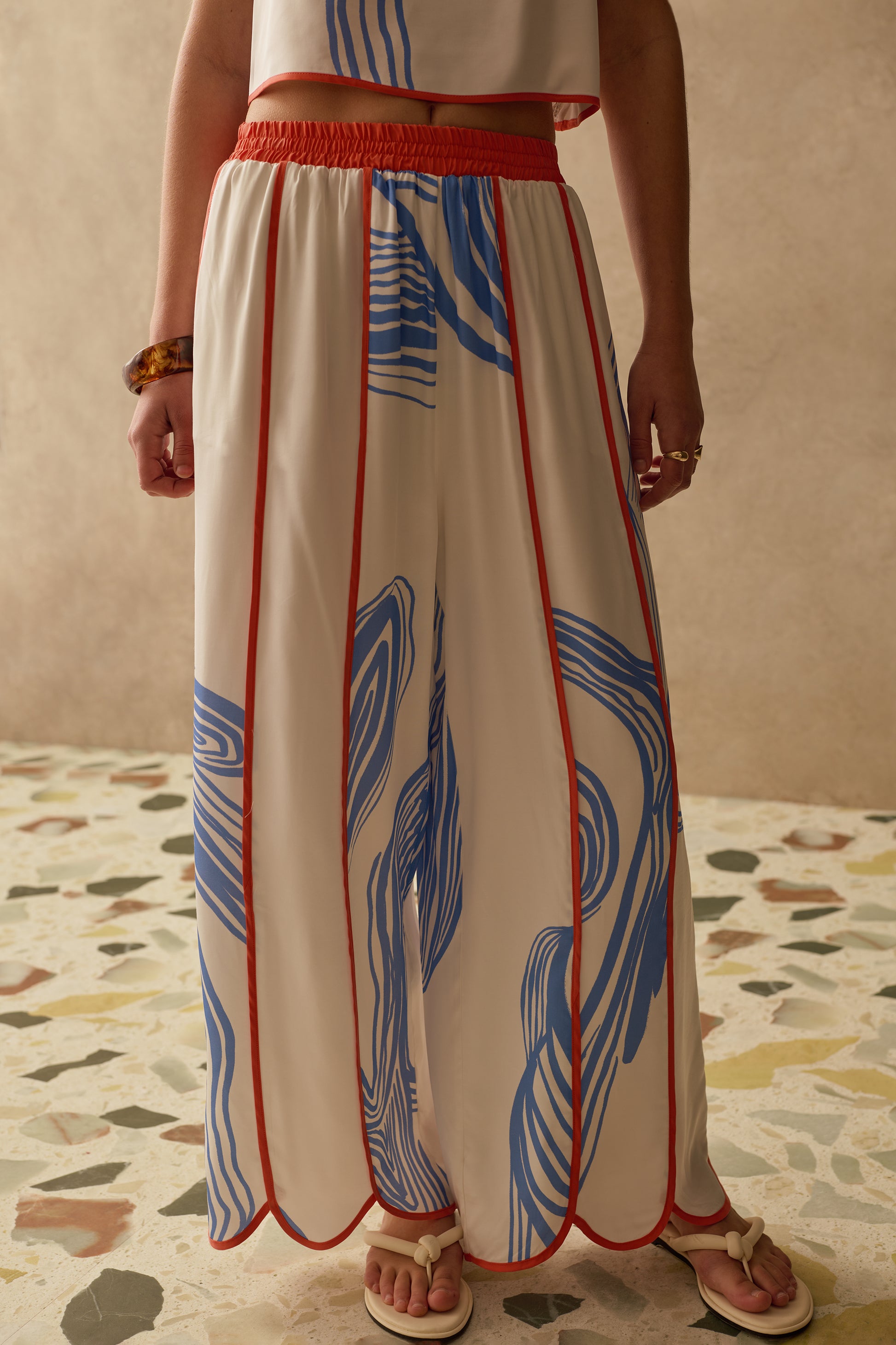 Person wearing wide leg patterned trousers with an orange waistband on a textured floor.