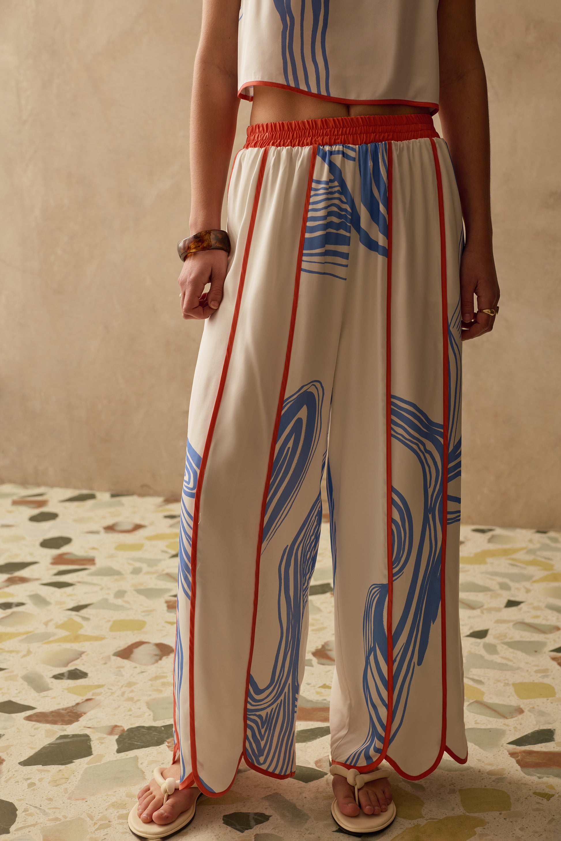 Person wearing a white wide leg trousers with blue and red patterns on a terracotta floor.