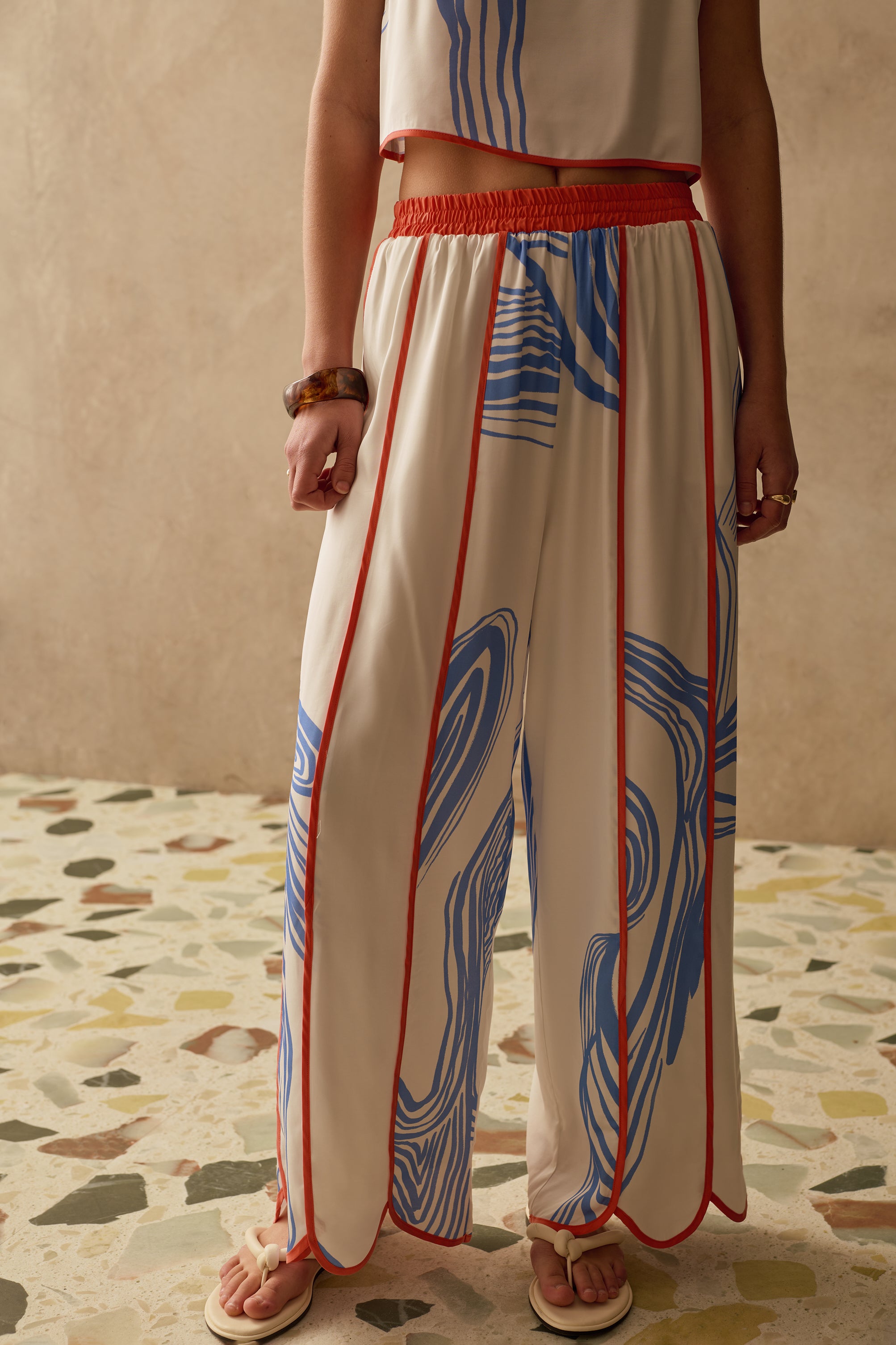 Person wearing a white wide leg trousers with blue and red patterns on a terracotta floor.