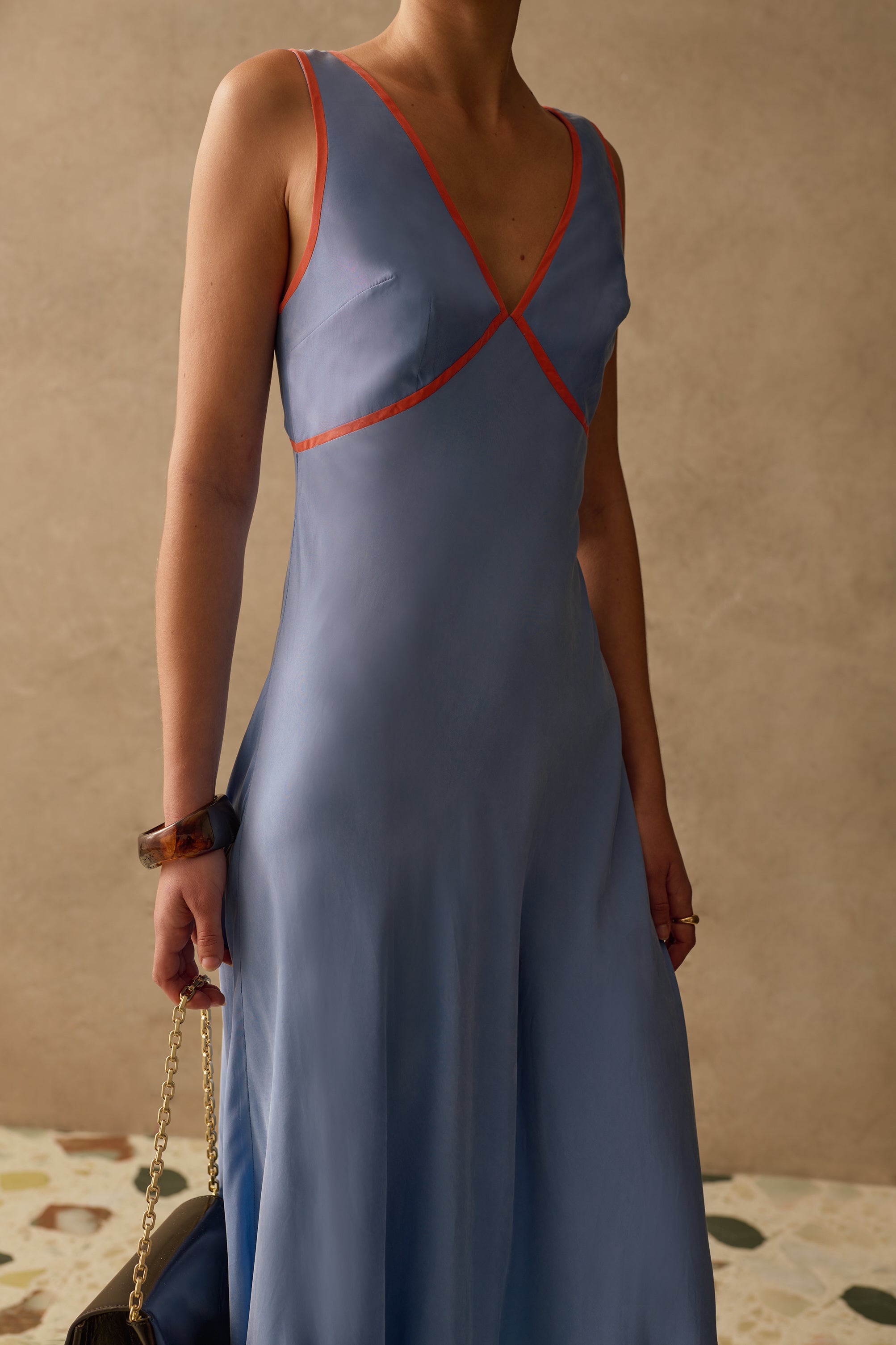 Woman wearing a blue v-neck sleeveless dress with red accents against a beige wall.