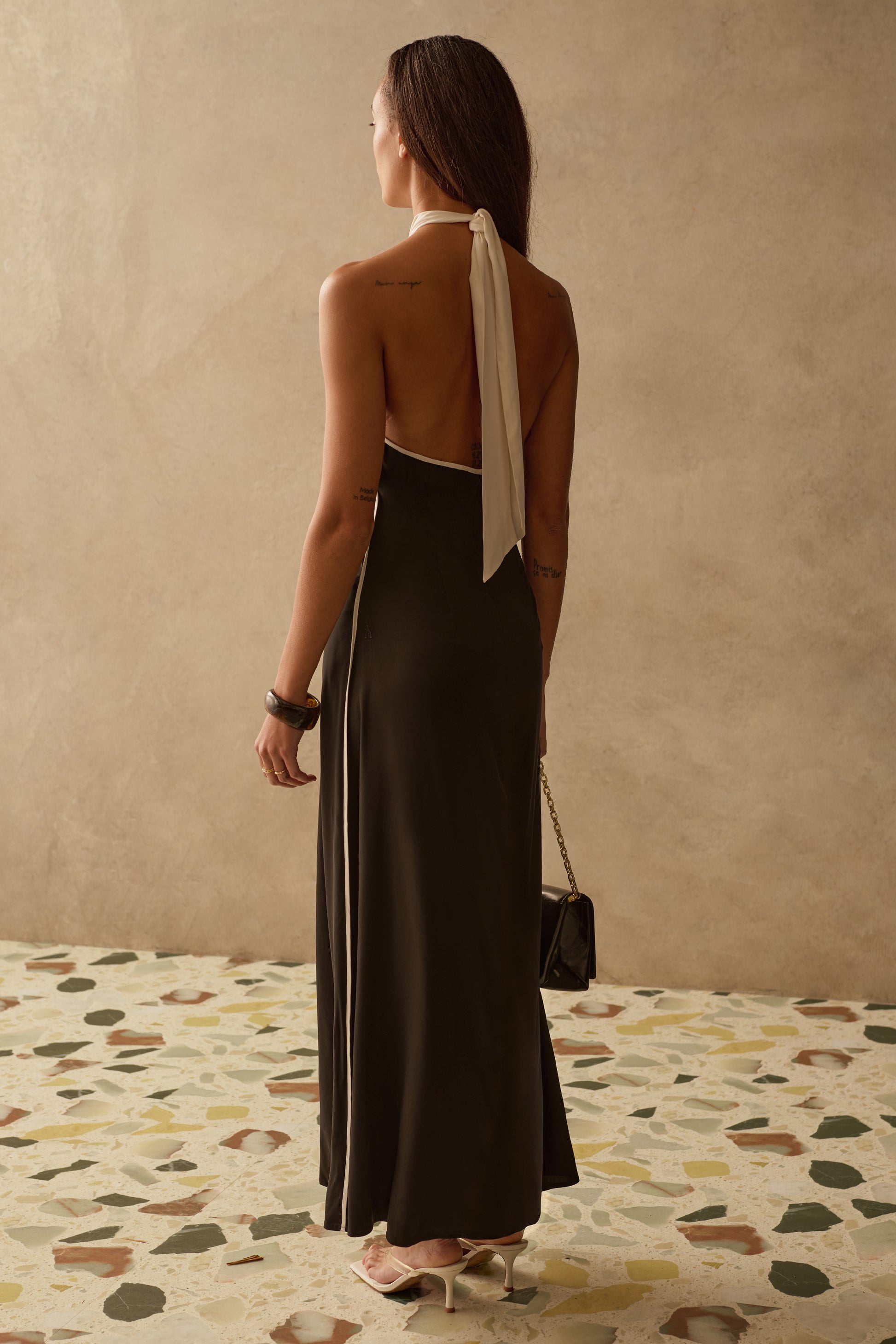 Model wearing a Sancia halter maxi dress holding a black bag and wearing white heels