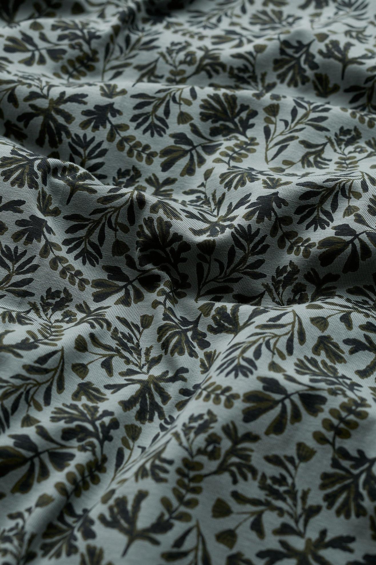 A close-up of draped fabric with an all-over botanical leaf pattern in dark olive green and black on a muted light gray background.