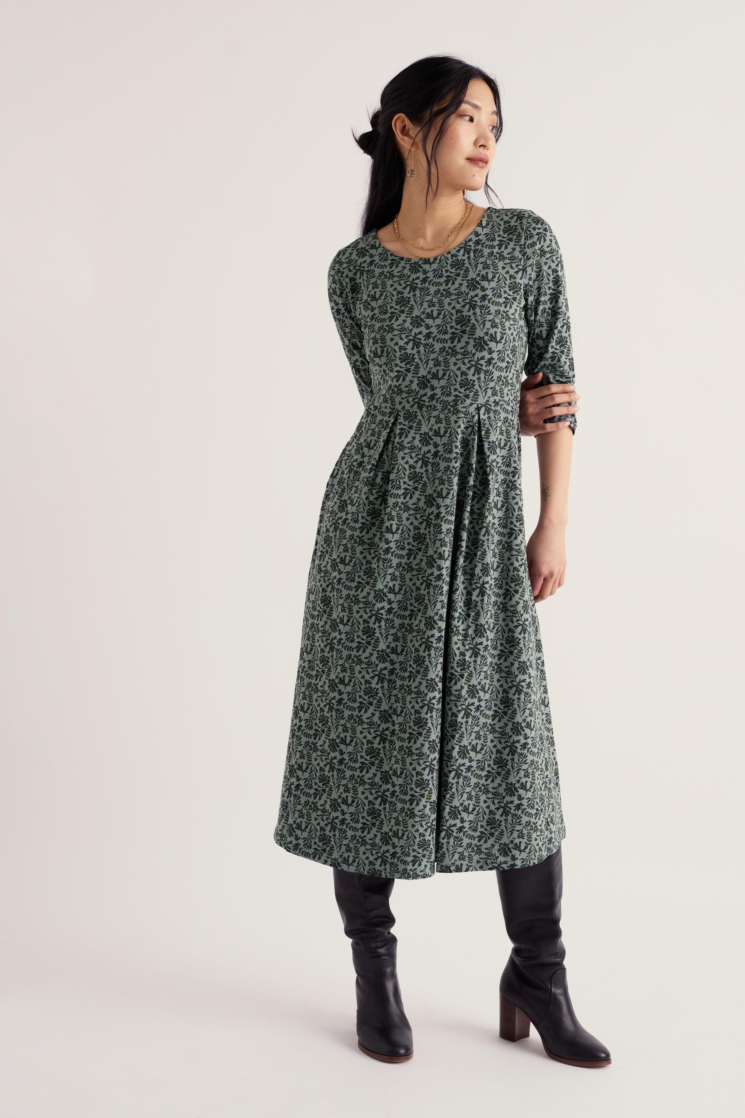 A woman modeling a dark green and black floral print midi dress with elbow-length sleeves, paired with black knee-high boots, against a plain light gray studio background.