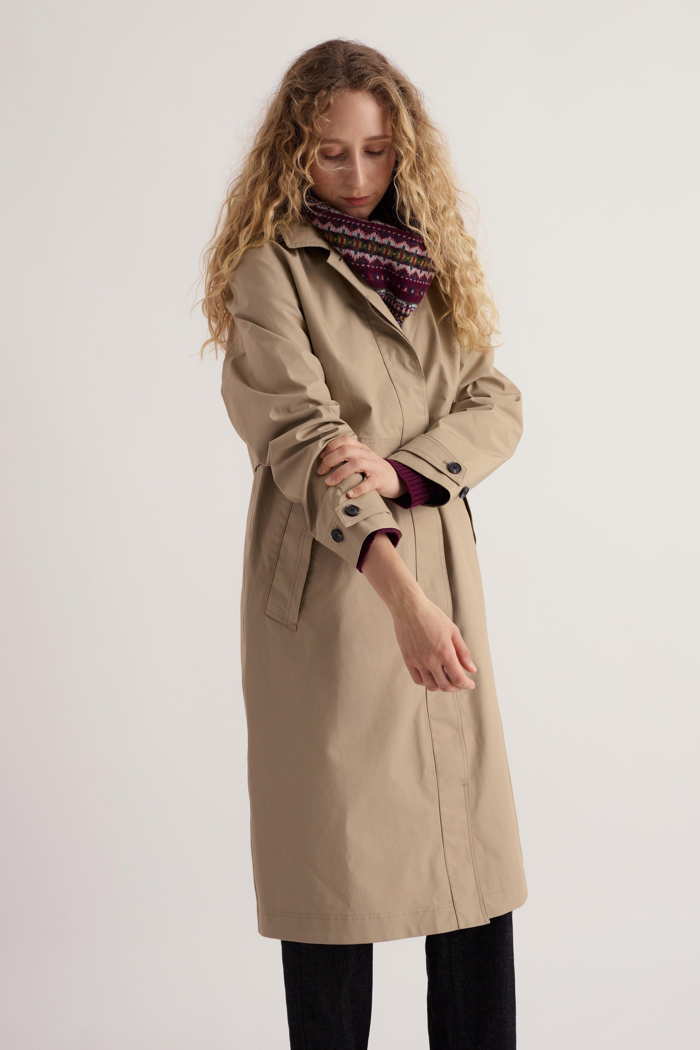 Woman wearing a beige Seasalt Cornwall trench coat over a colorful patterned scarf and knit sweater