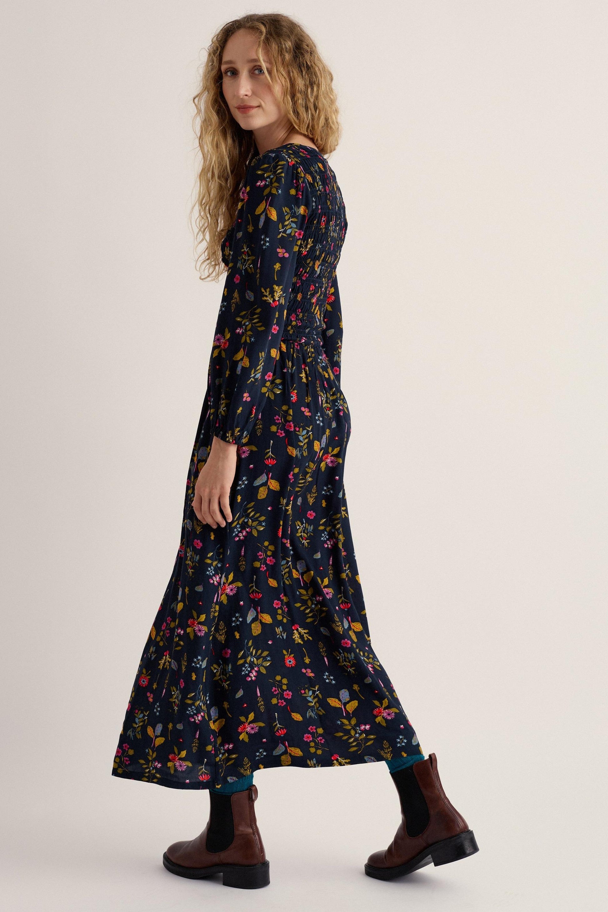 A woman modeling a long-sleeved navy maxi dress with a colorful floral print, paired with dark brown Chelsea boots, photographed against a plain light beige studio background.
