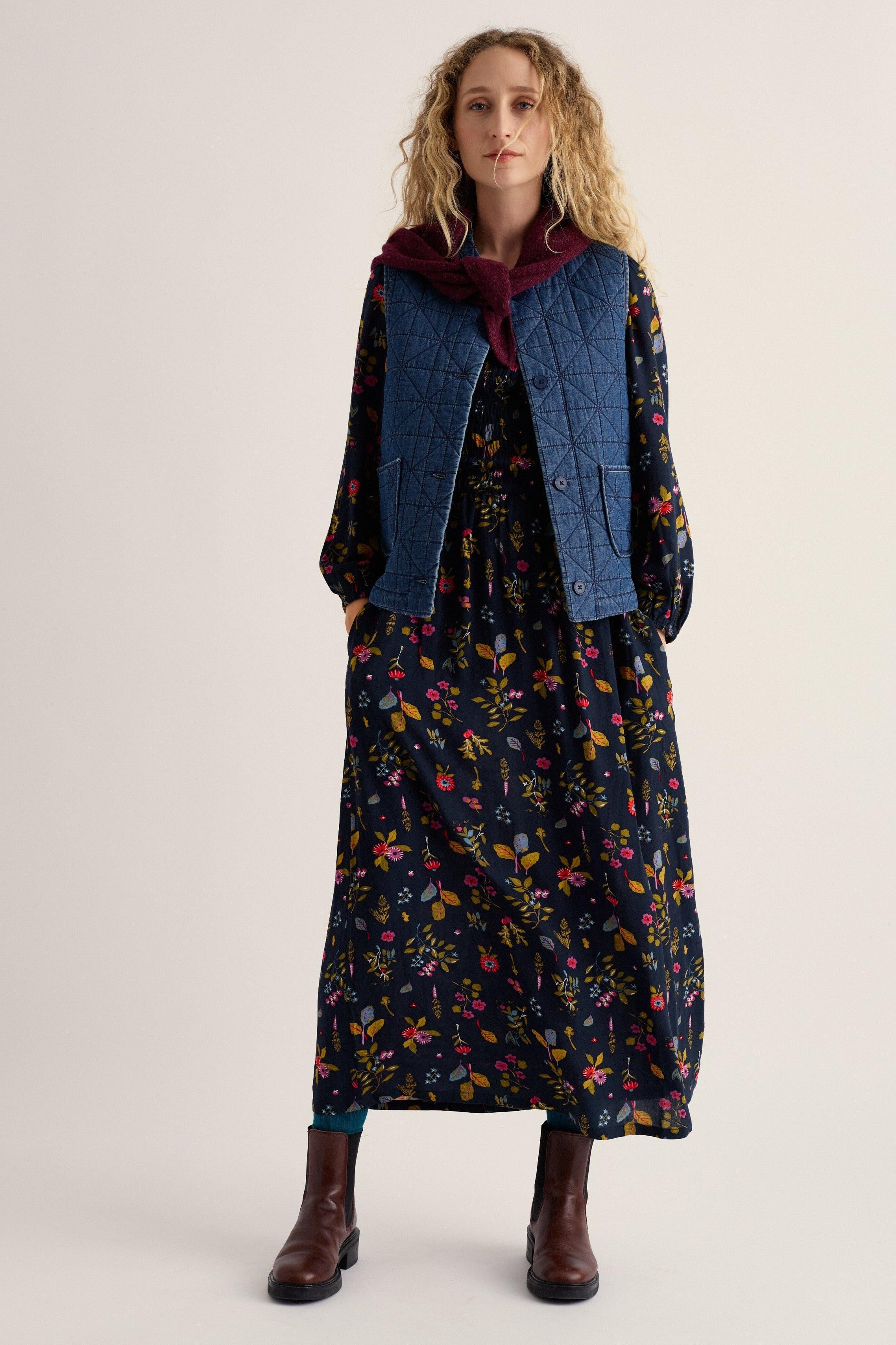 A woman stands against a plain light beige studio background wearing a long-sleeved dark floral dress with a blue quilted vest layered over it, a burgundy hood, and brown ankle boots.