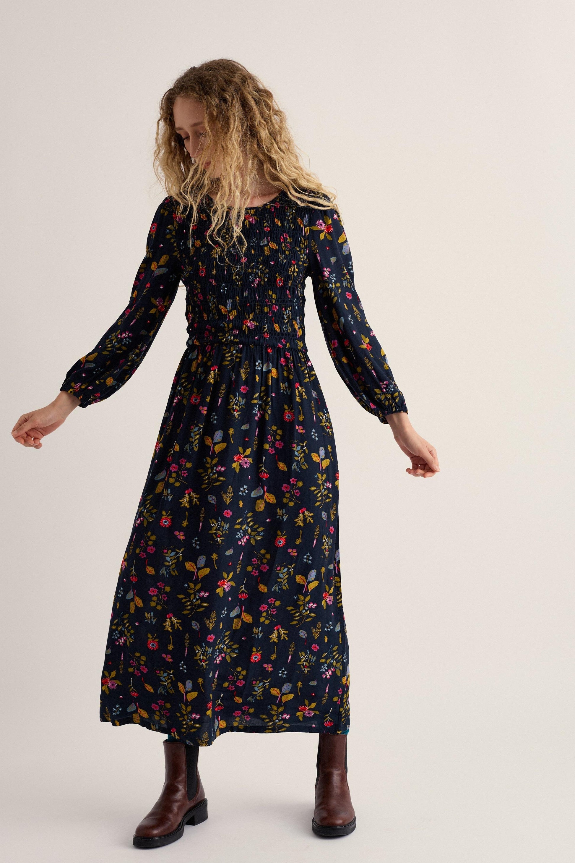 A woman modeling a long-sleeved midi dress with a dark navy blue background and a colorful floral print, paired with brown leather boots, against a plain light beige studio background.