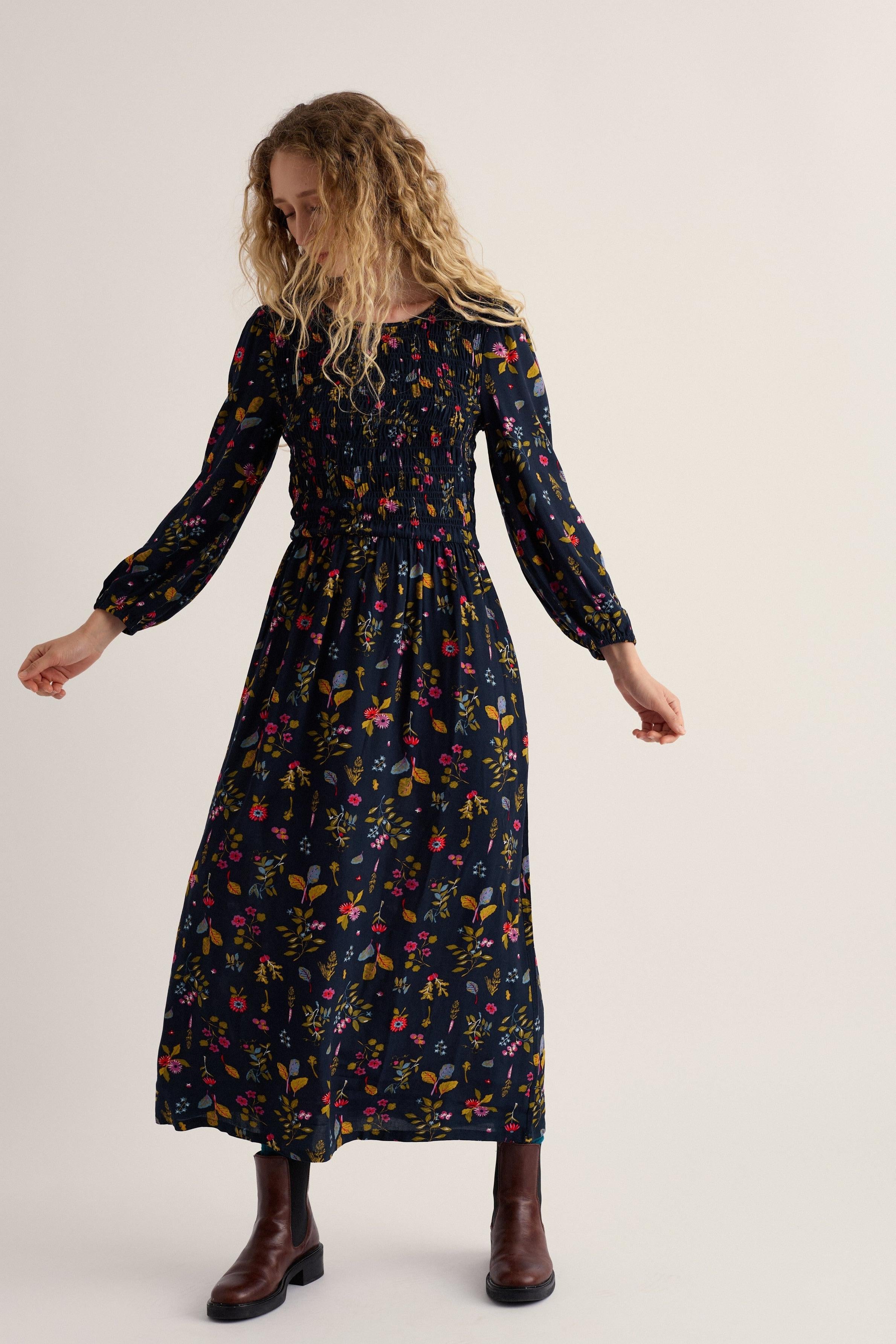A woman modeling a long-sleeved midi dress with a dark navy blue background and a colorful floral print, paired with brown leather boots, against a plain light beige studio background.