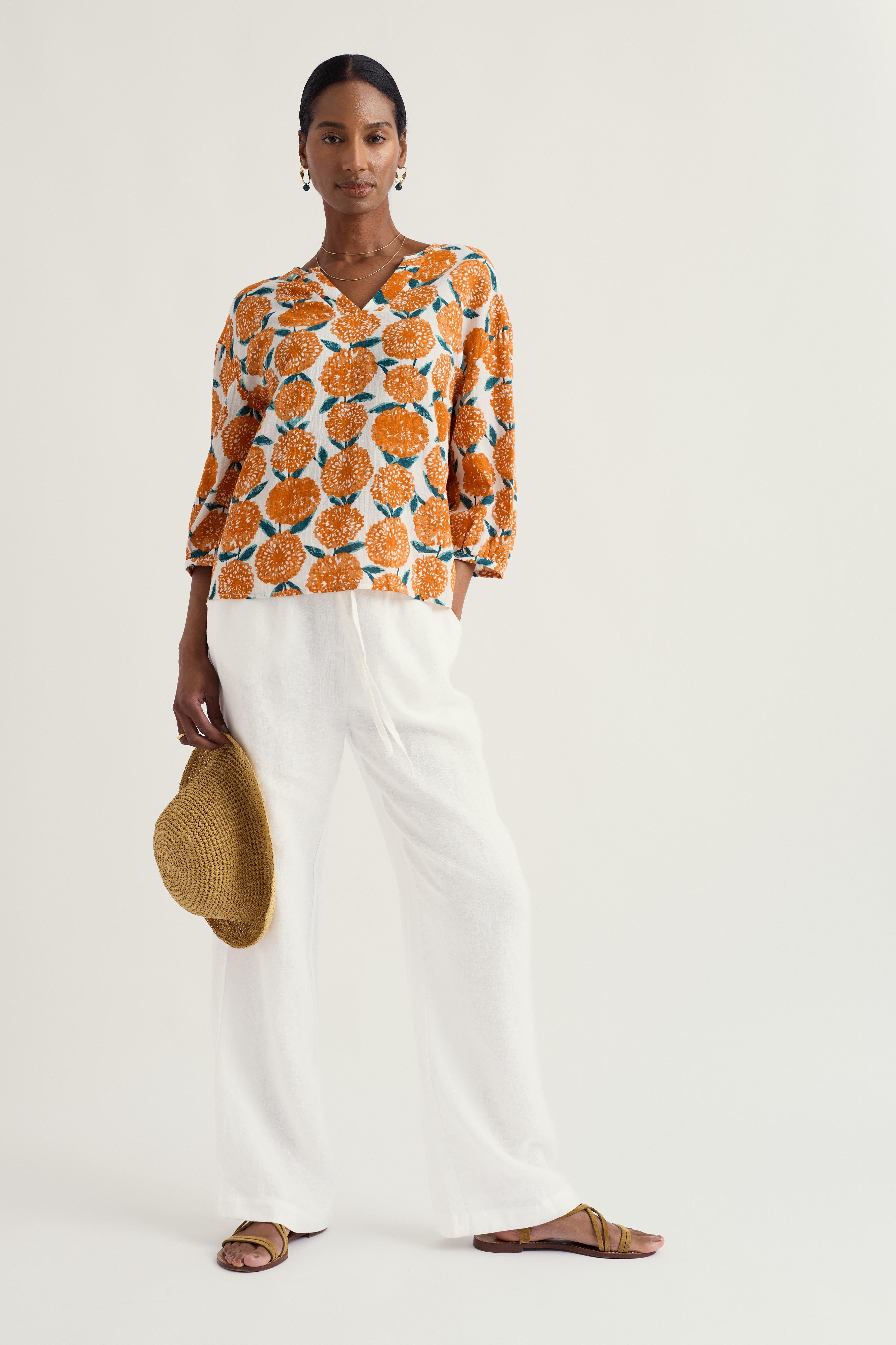 Woman wearing a patterned Seasalt top and white pants holding a straw hat on a plain background