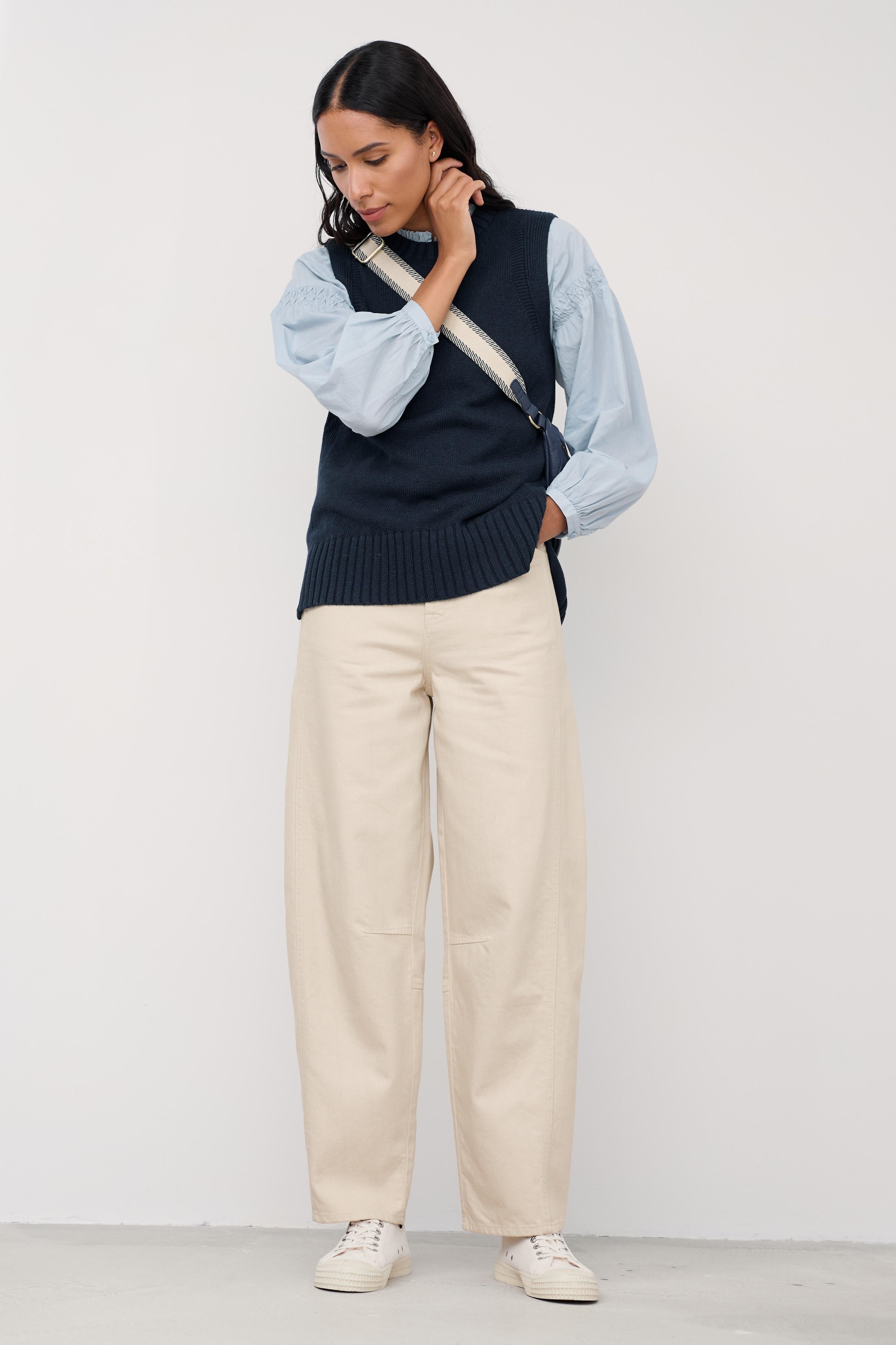Woman wearing a navy sweater vest, light blue shirt, beige organic cotton pants, and white sneakers on a white background