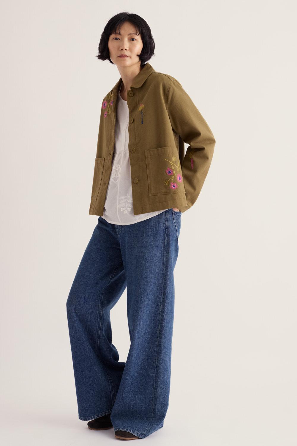 A woman wearing a khaki Seasalt organic cotton jacket with floral embroidery over a white top and blue wide-leg jeans.
