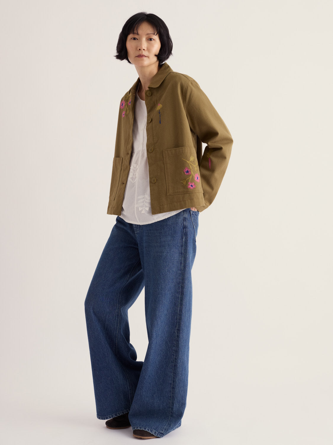 A woman wearing a khaki Seasalt organic cotton jacket with floral embroidery over a white top and blue wide-leg jeans.
