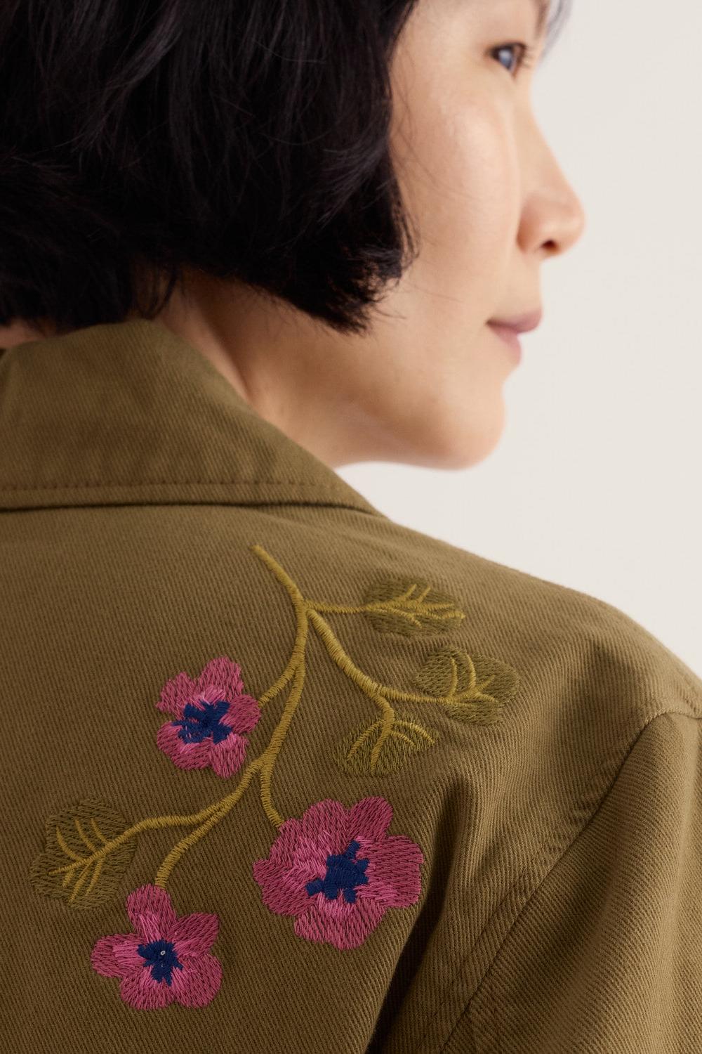 A woman wearing a khaki Seasalt jacket with detailed floral embroidery on the shoulder.