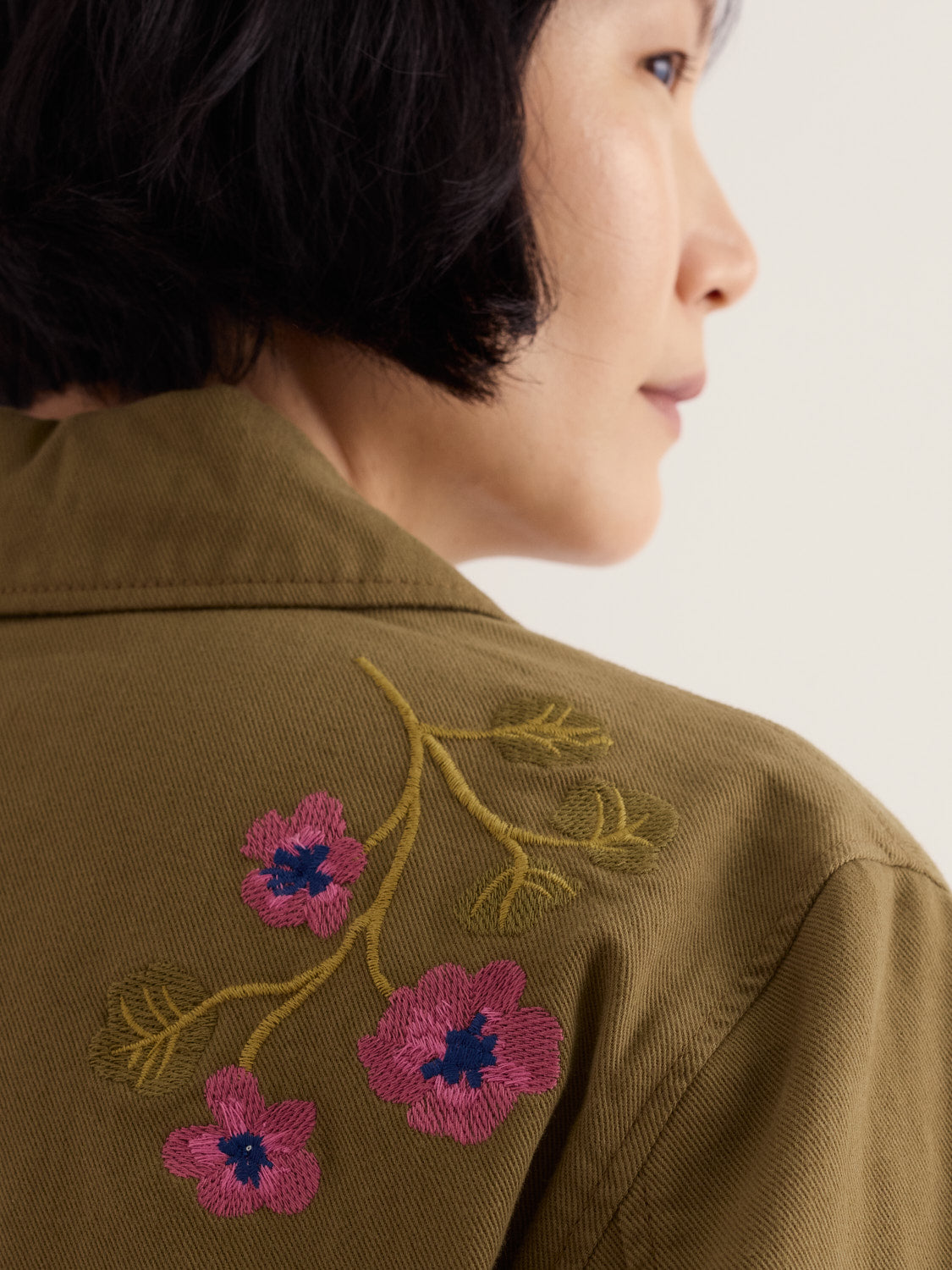 A woman wearing a khaki Seasalt jacket with detailed floral embroidery on the shoulder.