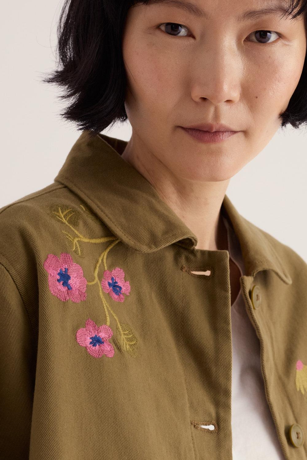 Close-up of a woman wearing a khaki Seasalt jacket featuring detailed floral embroidery on the shoulder.