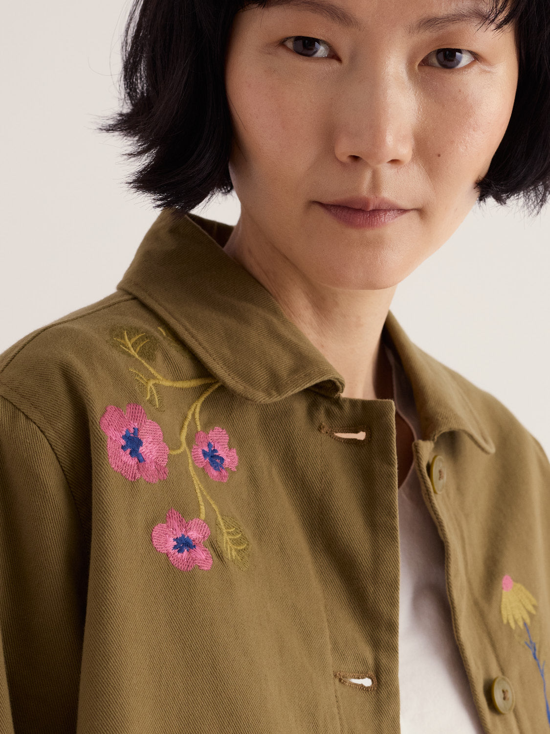 Close-up of a woman wearing a khaki Seasalt jacket featuring detailed floral embroidery on the shoulder.