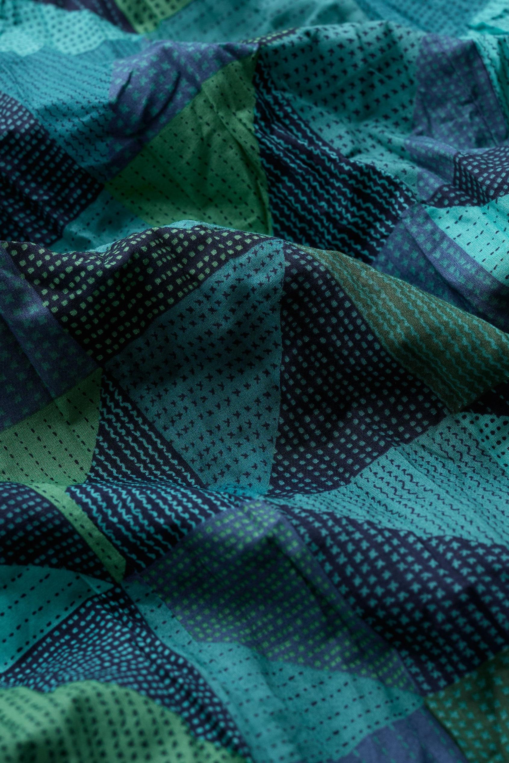 A close-up of fabric with a geometric patchwork pattern in shades of teal, green, and dark navy blue, featuring dotted and dashed triangle motifs.