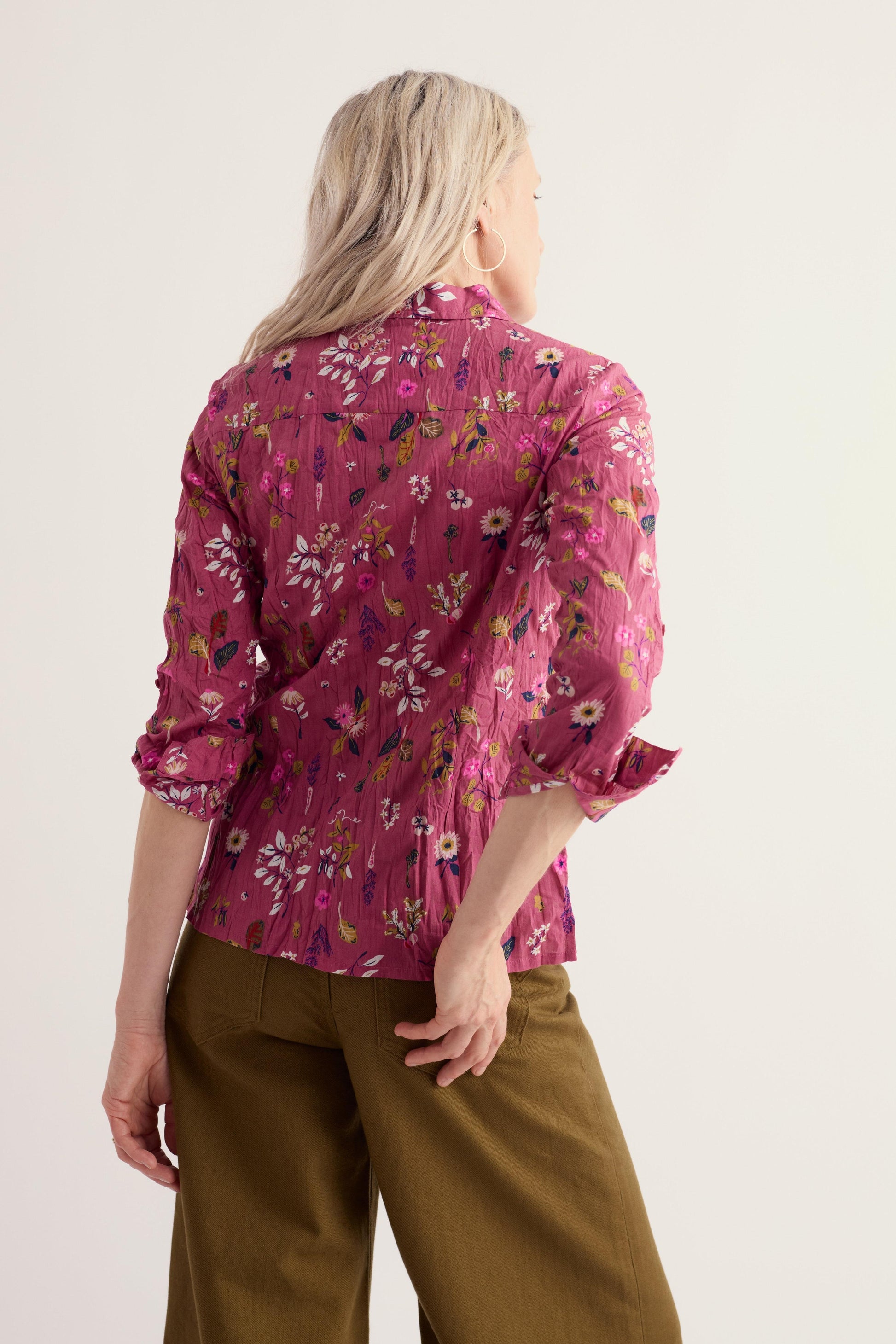 A woman shown from the back wearing a pink floral long-sleeve blouse with pleated detailing and olive green trousers, against a plain light background.