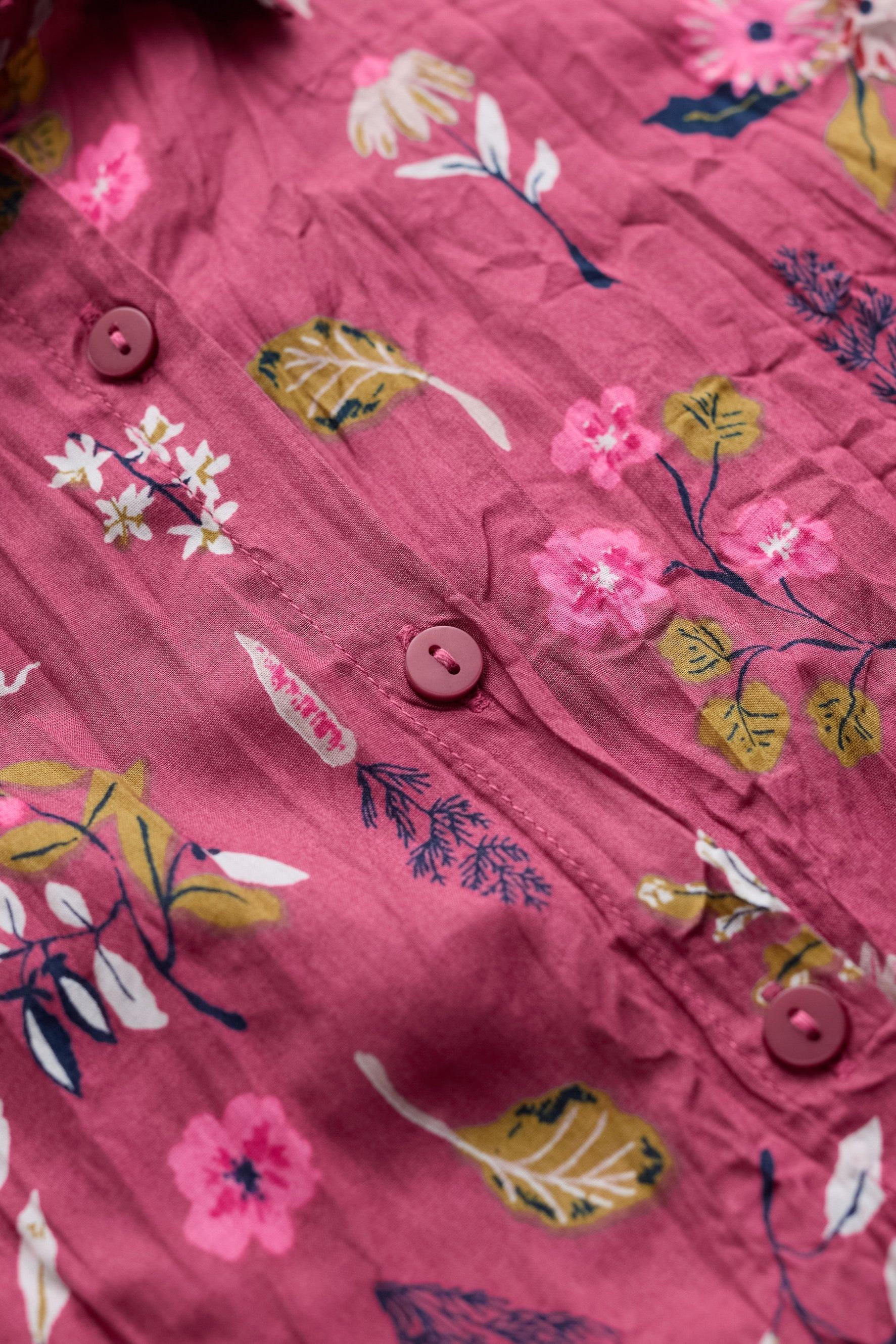 A close-up of a pink crinkled fabric garment with a floral print featuring pink flowers, green and yellow leaves, and brown buttons.