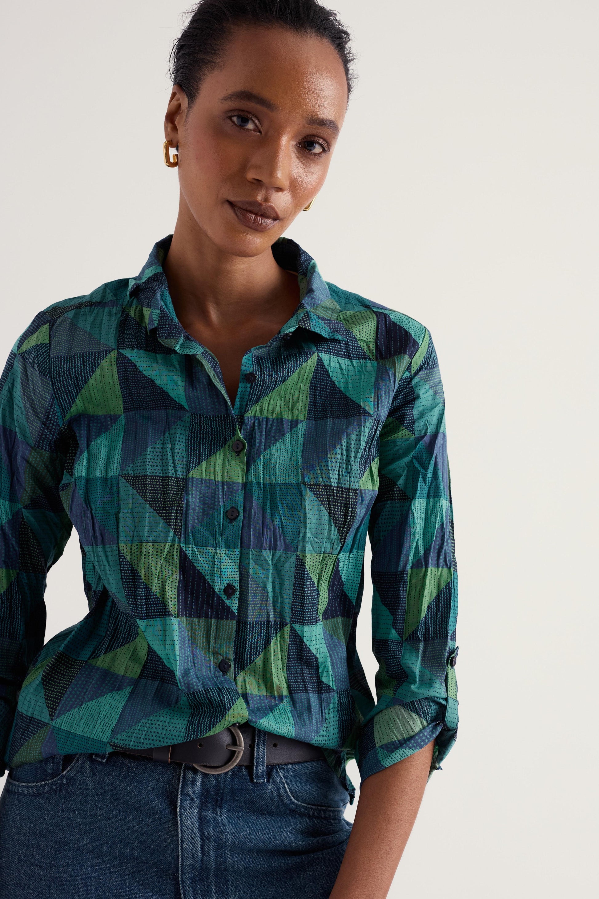 A woman wearing a long-sleeve button-up shirt with a geometric green, teal, and navy pattern, paired with dark blue jeans and a dark belt, against a plain light background.