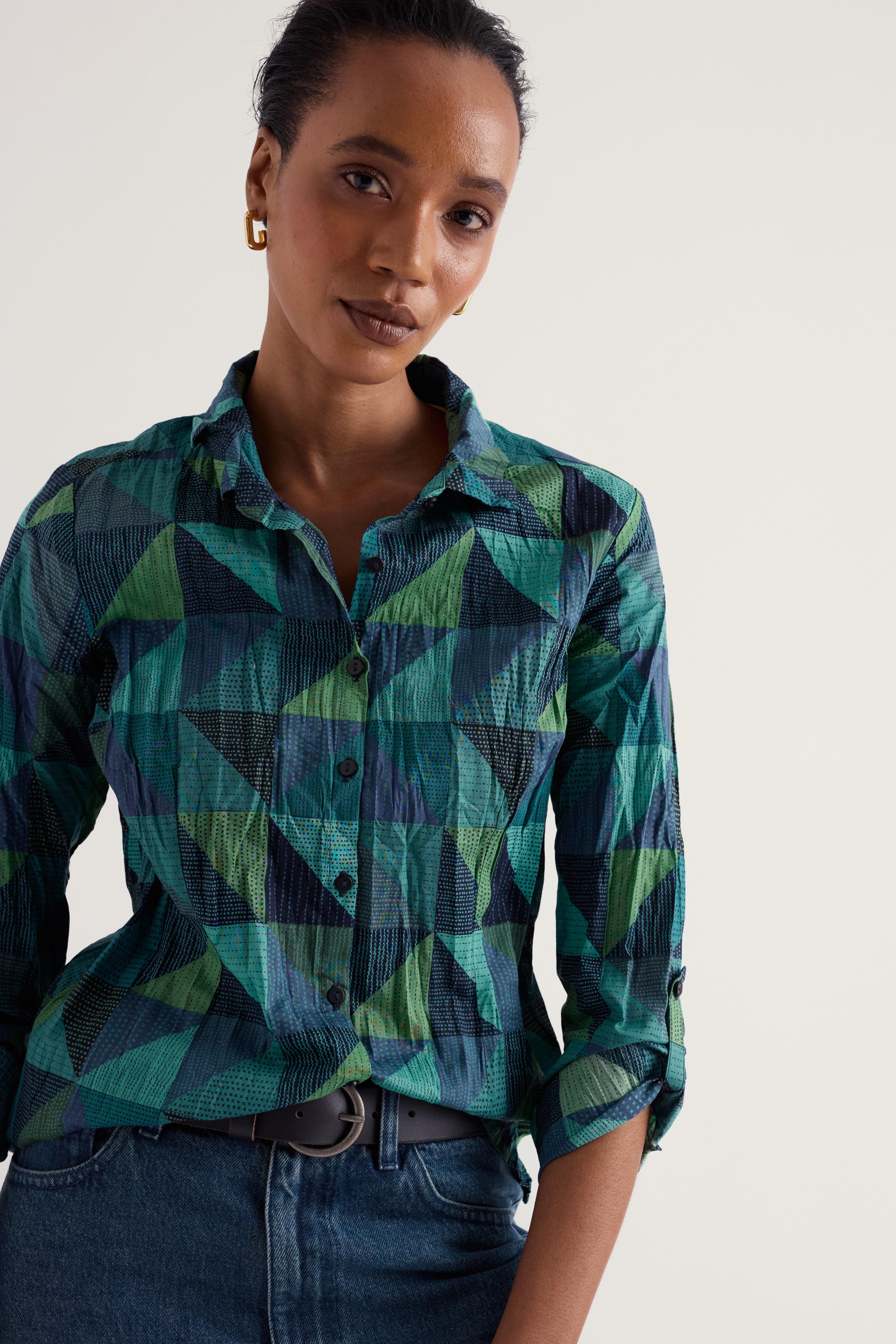 A woman wearing a long-sleeve button-up shirt with a geometric green, teal, and navy pattern, paired with dark blue jeans and a dark belt, against a plain light background.