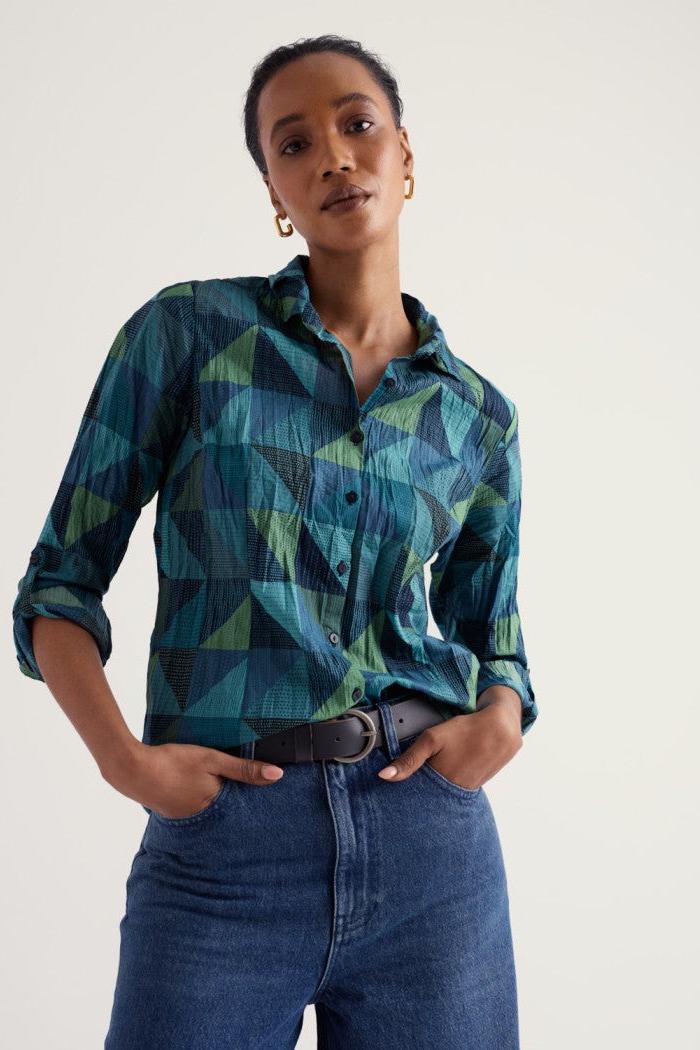 A woman wearing a long-sleeve button-up shirt with a blue, green, and teal geometric pattern, tucked into blue jeans with a dark belt, against a plain light background.