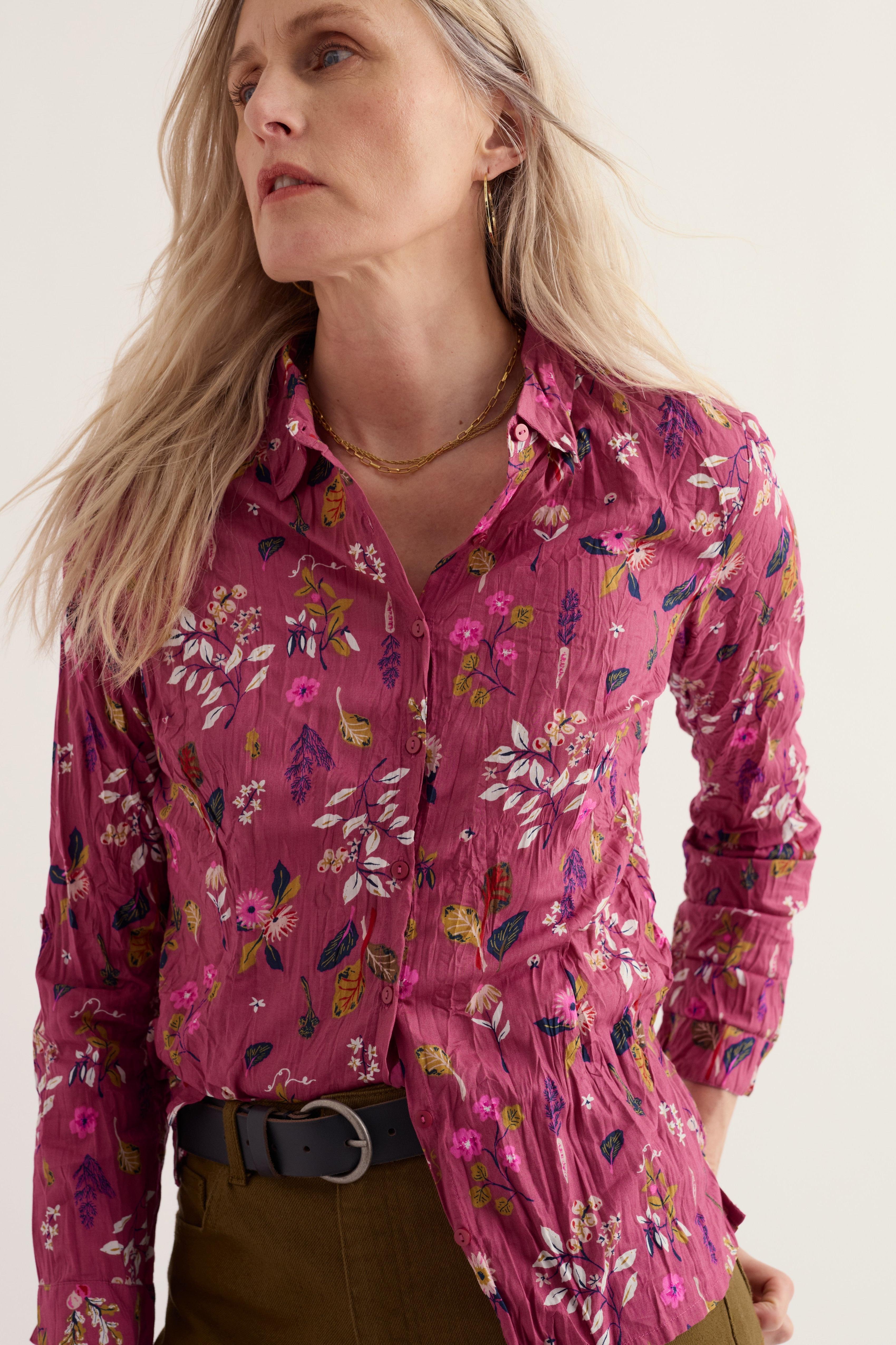A woman wearing a pink floral button-up shirt with a crinkled texture, paired with olive green pants and a black belt, against a plain off-white background.