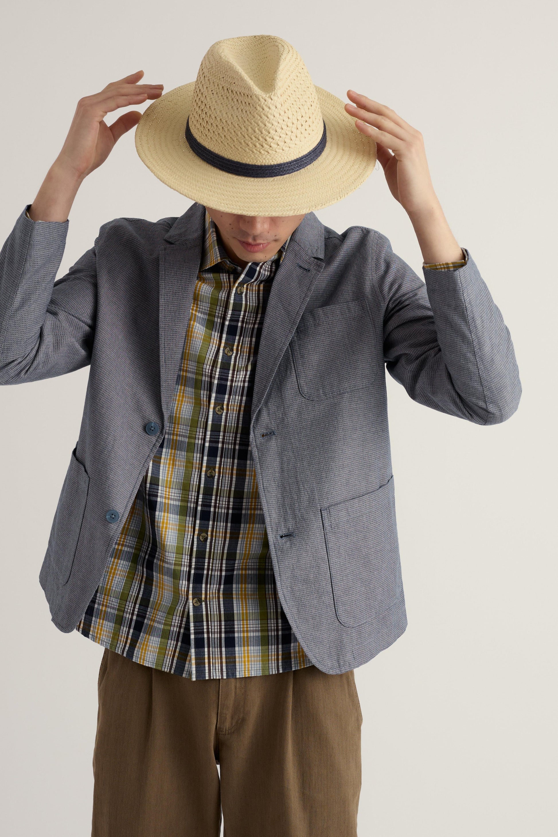 Person wearing a straw hat, gray organic cotton blazer, plaid shirt, and brown pants on a plain background