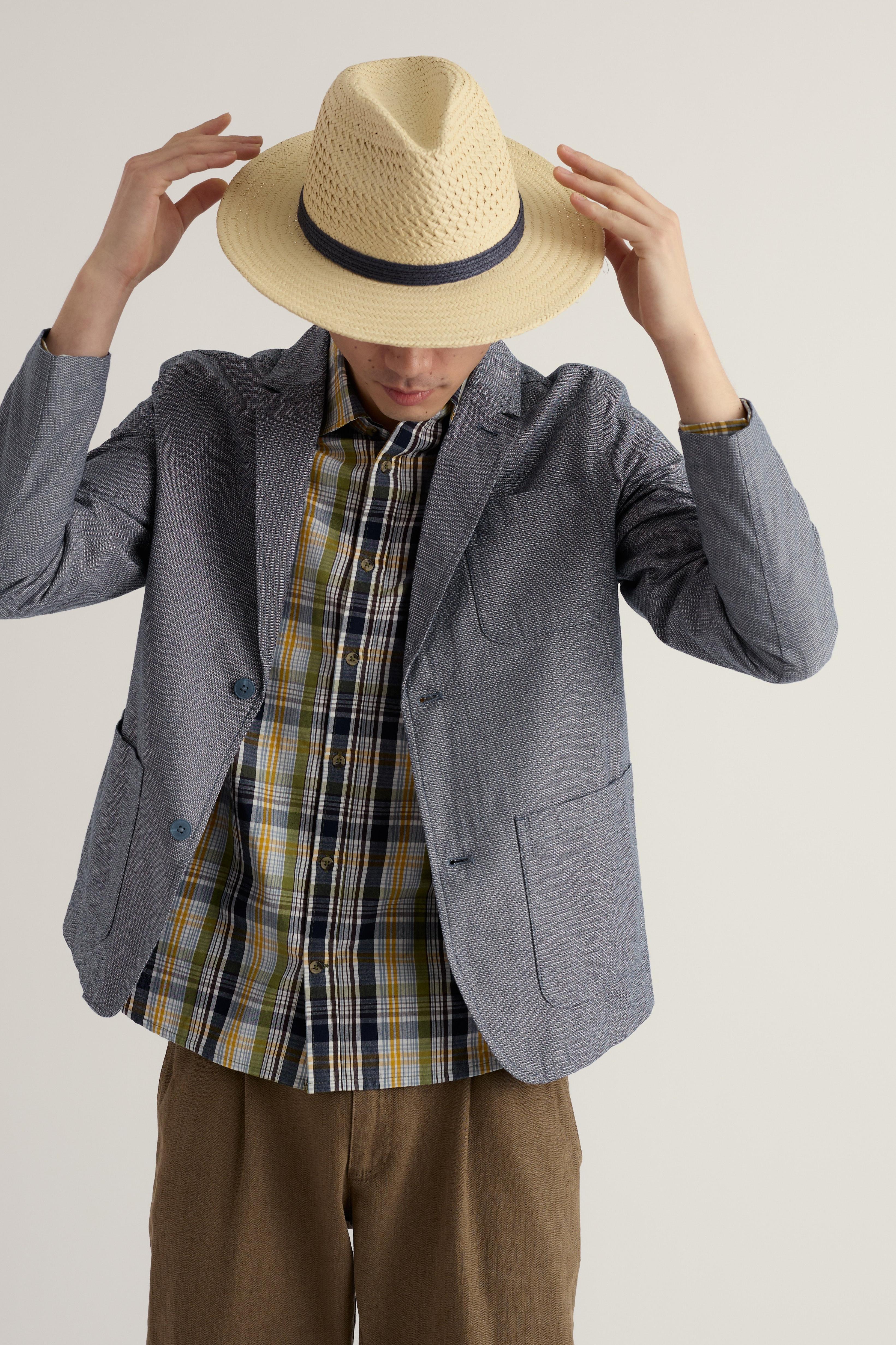 Person wearing a straw hat, gray organic cotton blazer, plaid shirt, and brown pants on a plain background
