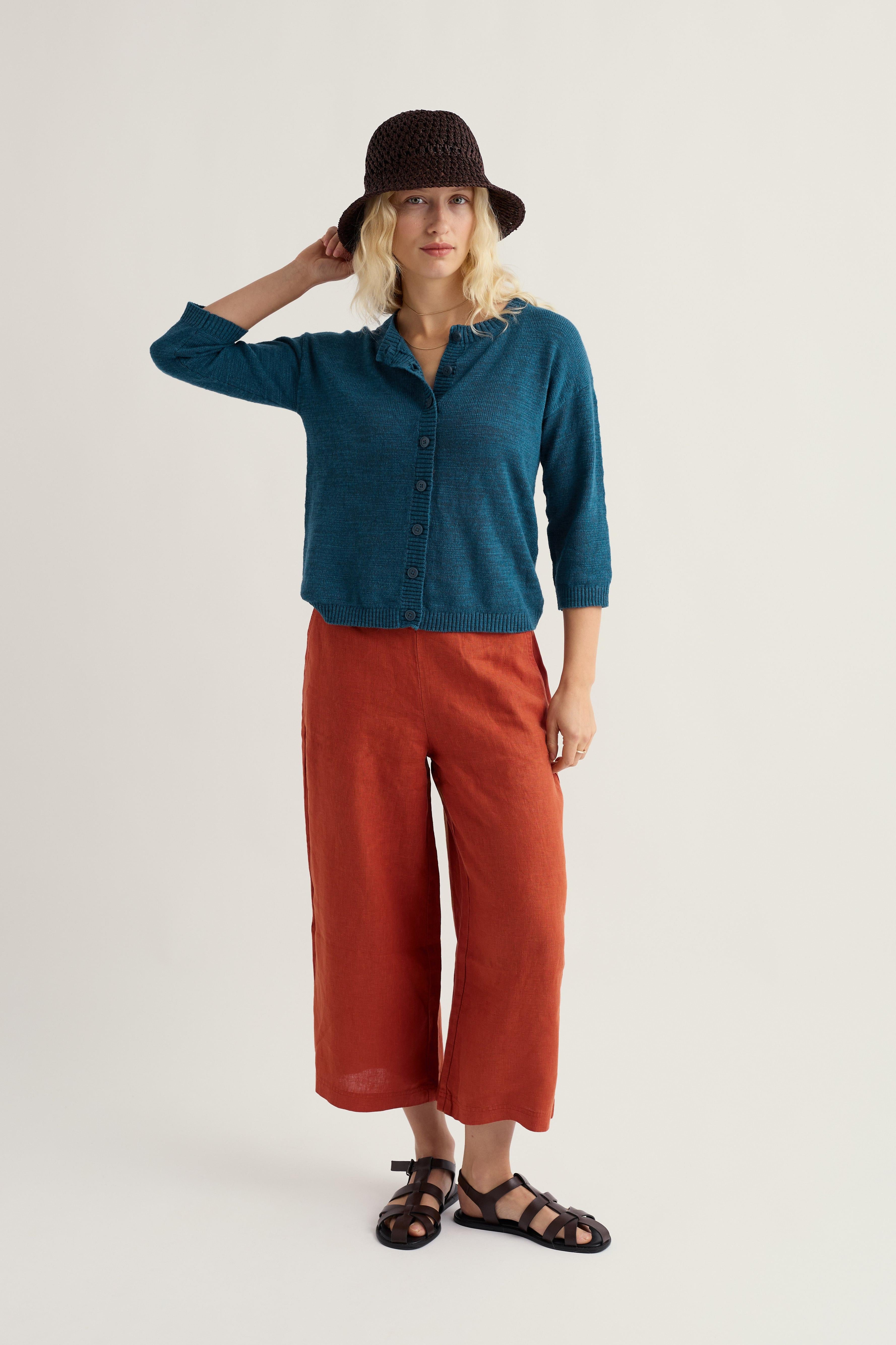 Woman wearing a teal organic cotton cardigan and rust-coloured pants with a black hat on a white background