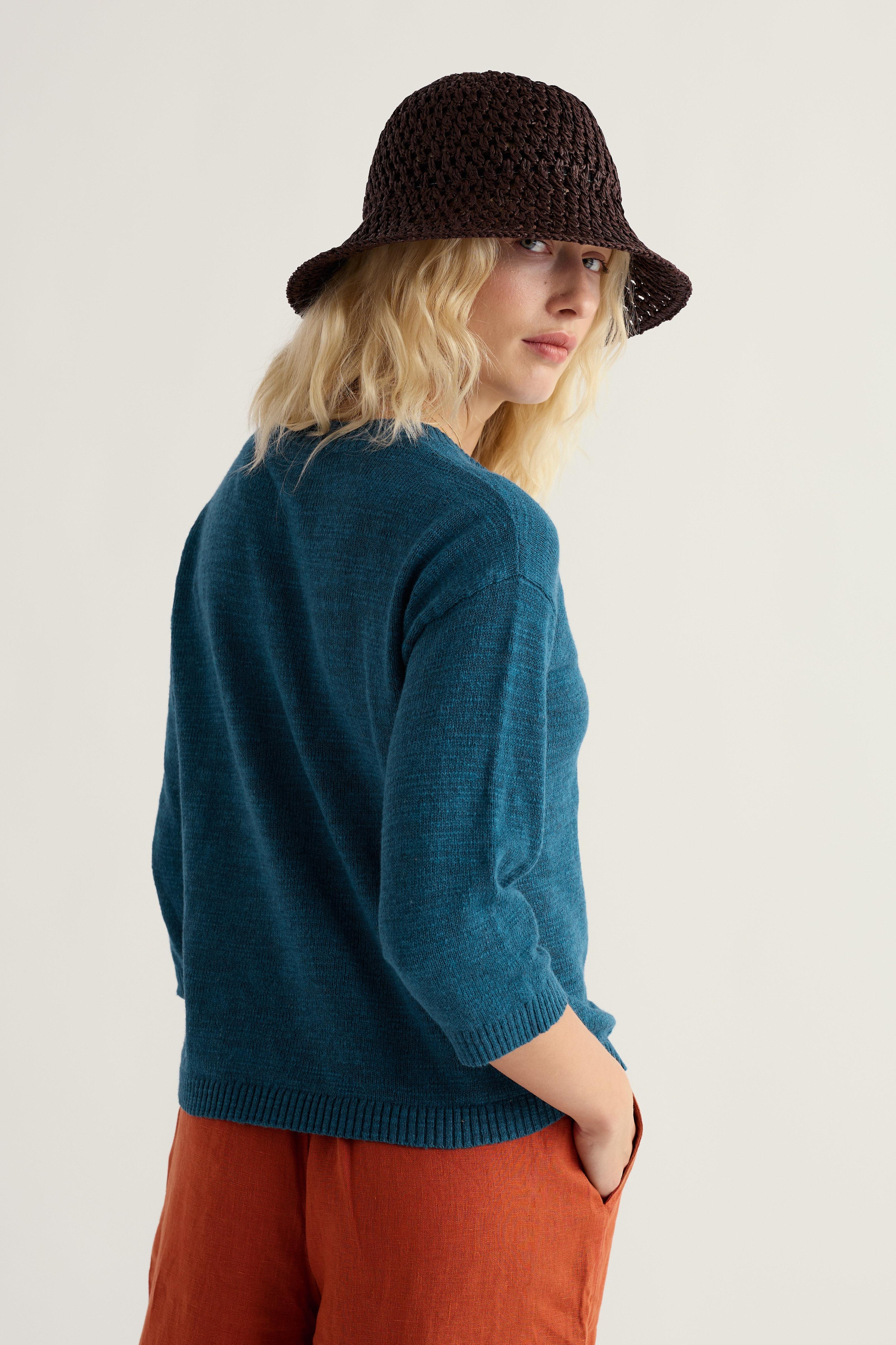Woman wearing a teal Seasalt cardigan and brown hat against a plain background