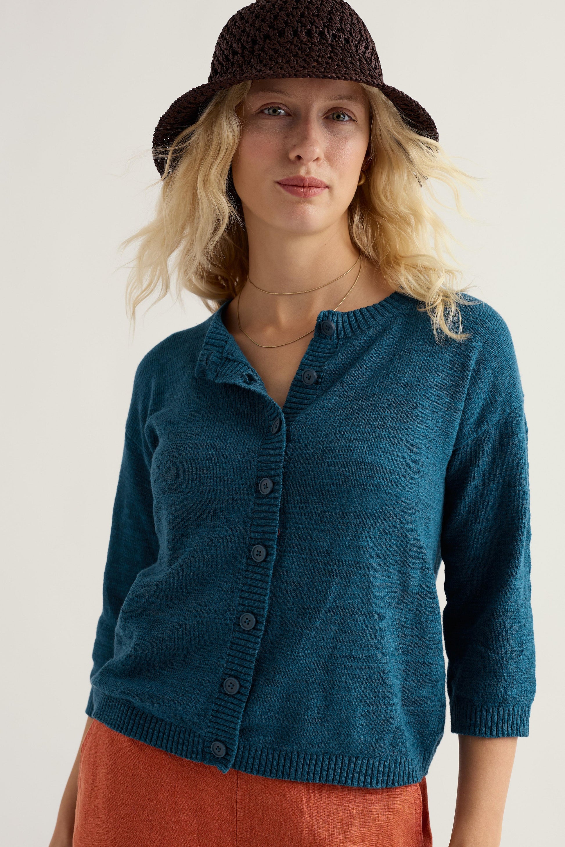 Woman wearing a teal organic cotton cardigan and brown hat against a white background